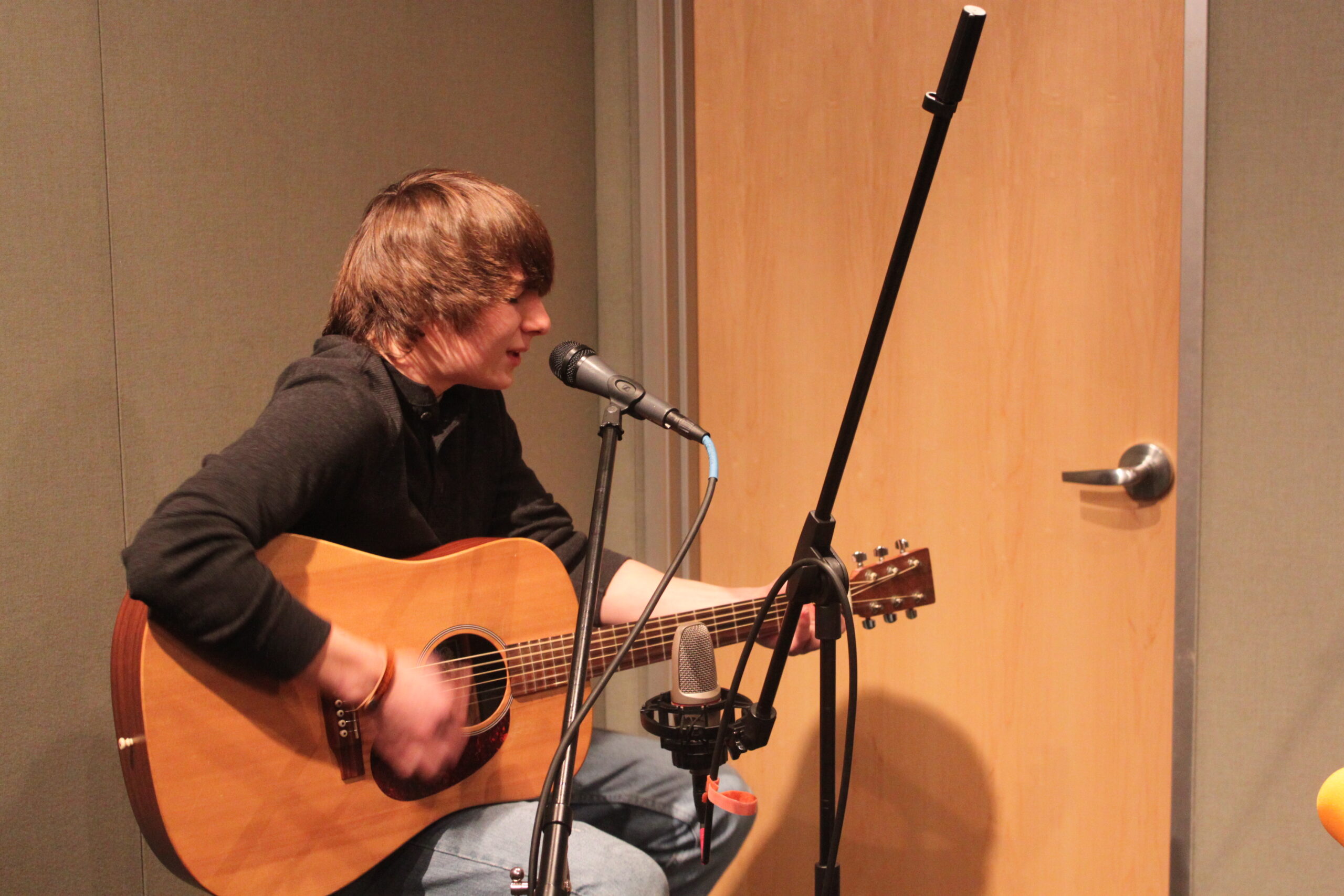 A teen sitting and playing acoustic guitar and singing into a microphone in a radio studio