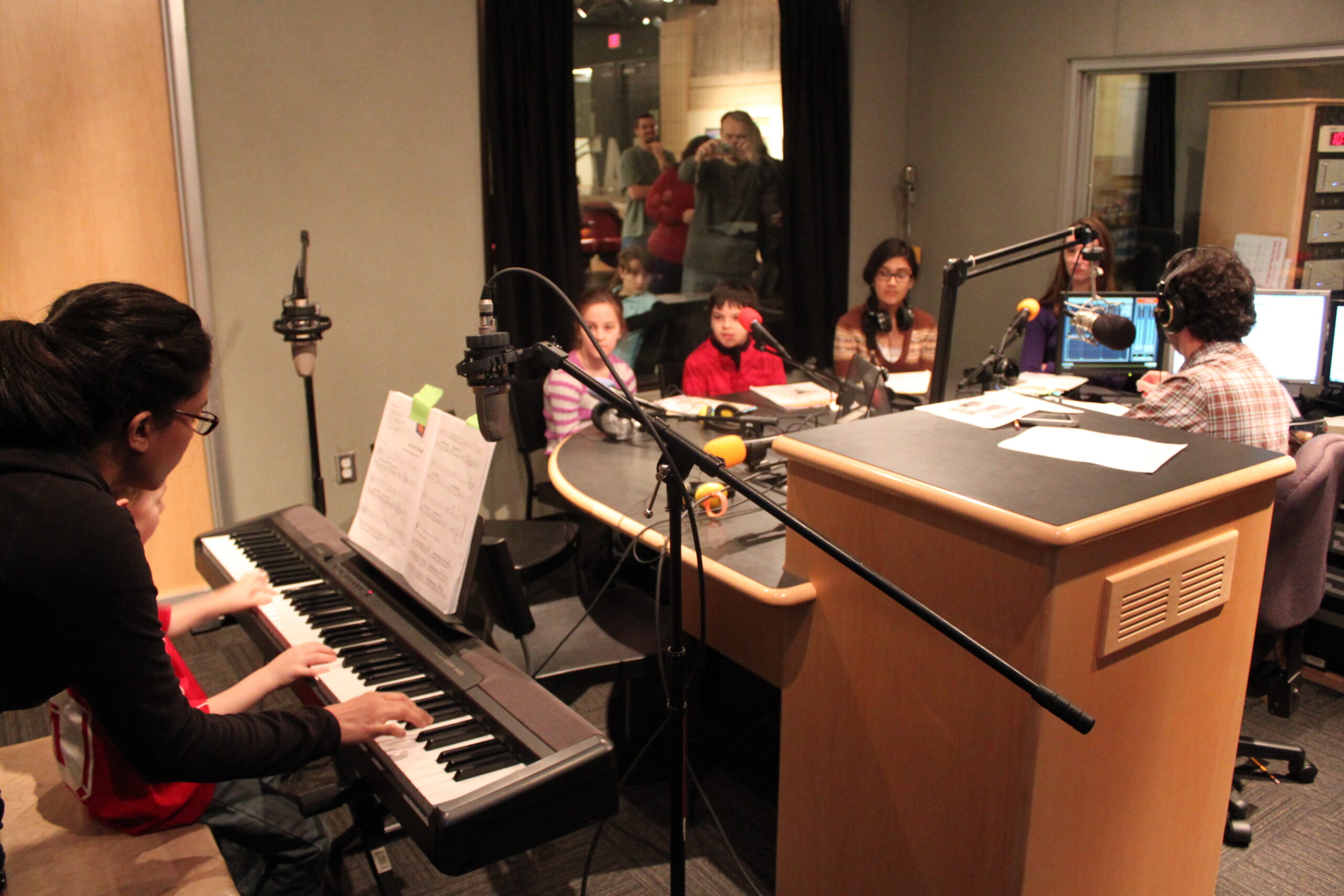 A youth playing a piano keyboard and four youth and one adult sitting behind microphones in a radio studio
