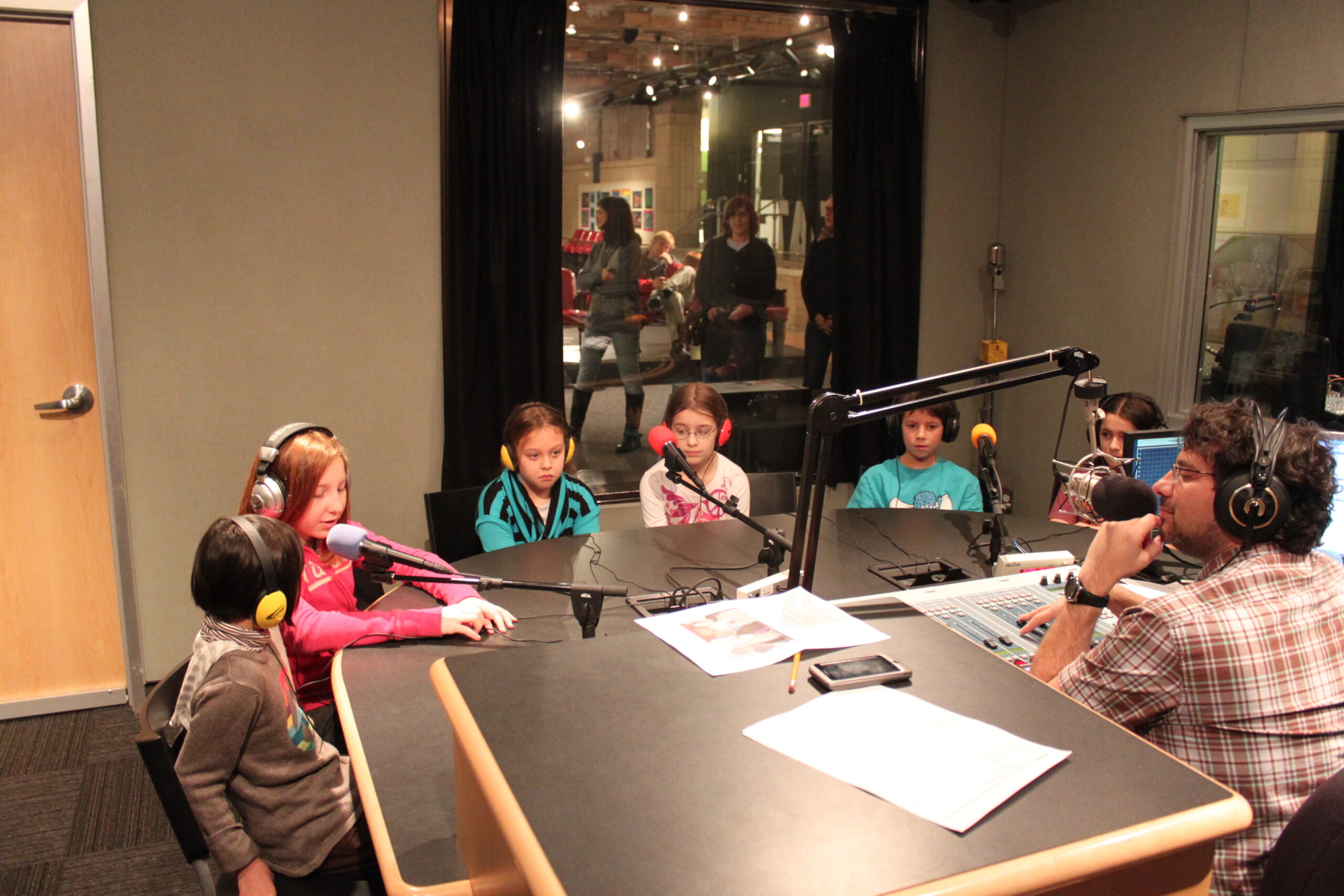 Six youth wearing headphones speaking into microphones being interviewed by an adult wearing headphones and sitting behind a microphone and an audio mixer in a radio studio