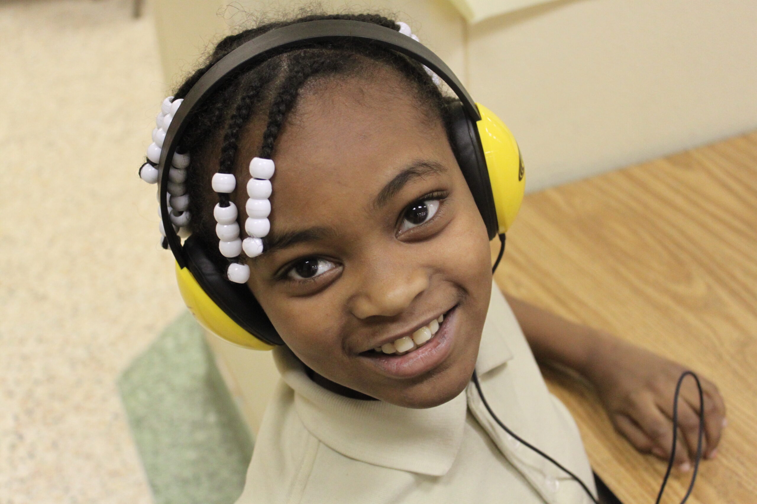 A youth wearing headphones sitting at a classroom desk and smiling up at the camera