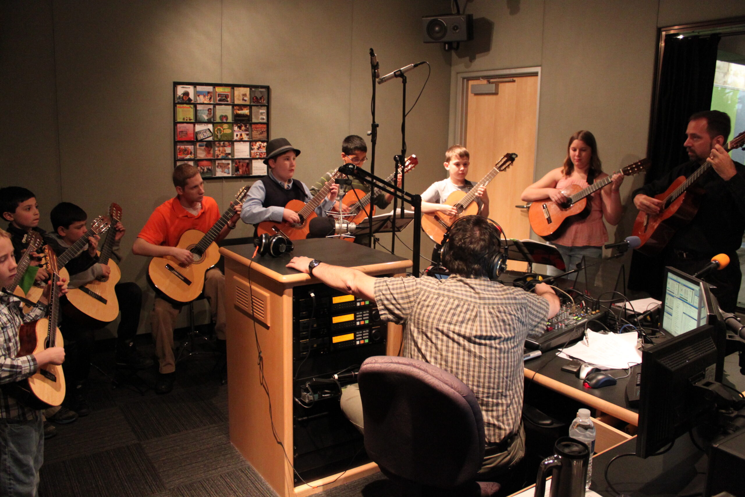 Eight youth and an adult playing acoustic guitars as an adult sitting behind a microphone and an audio mixer listens in a radio studio