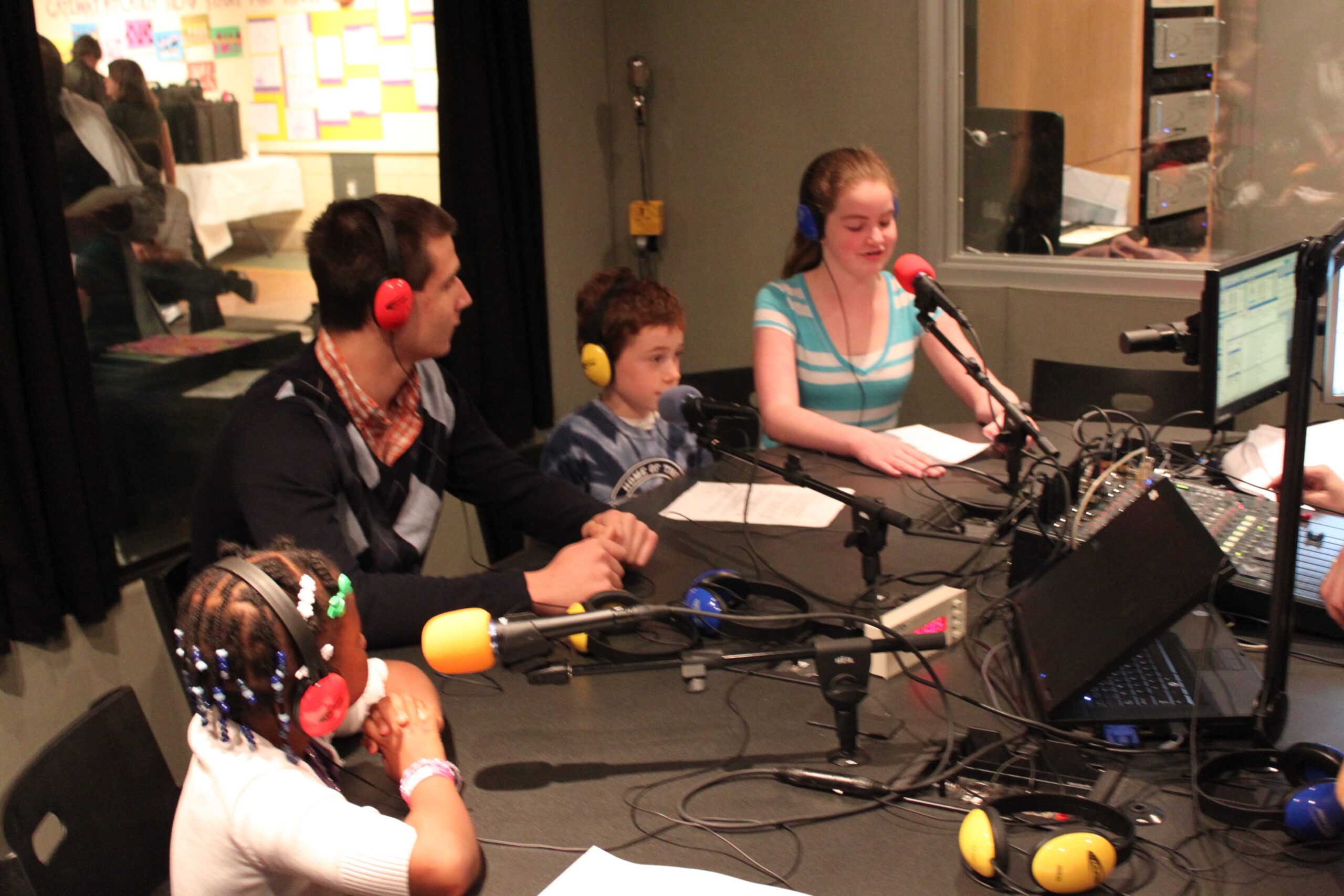 An adult and three youth wearing headphones speaking into microphones in a radio studio