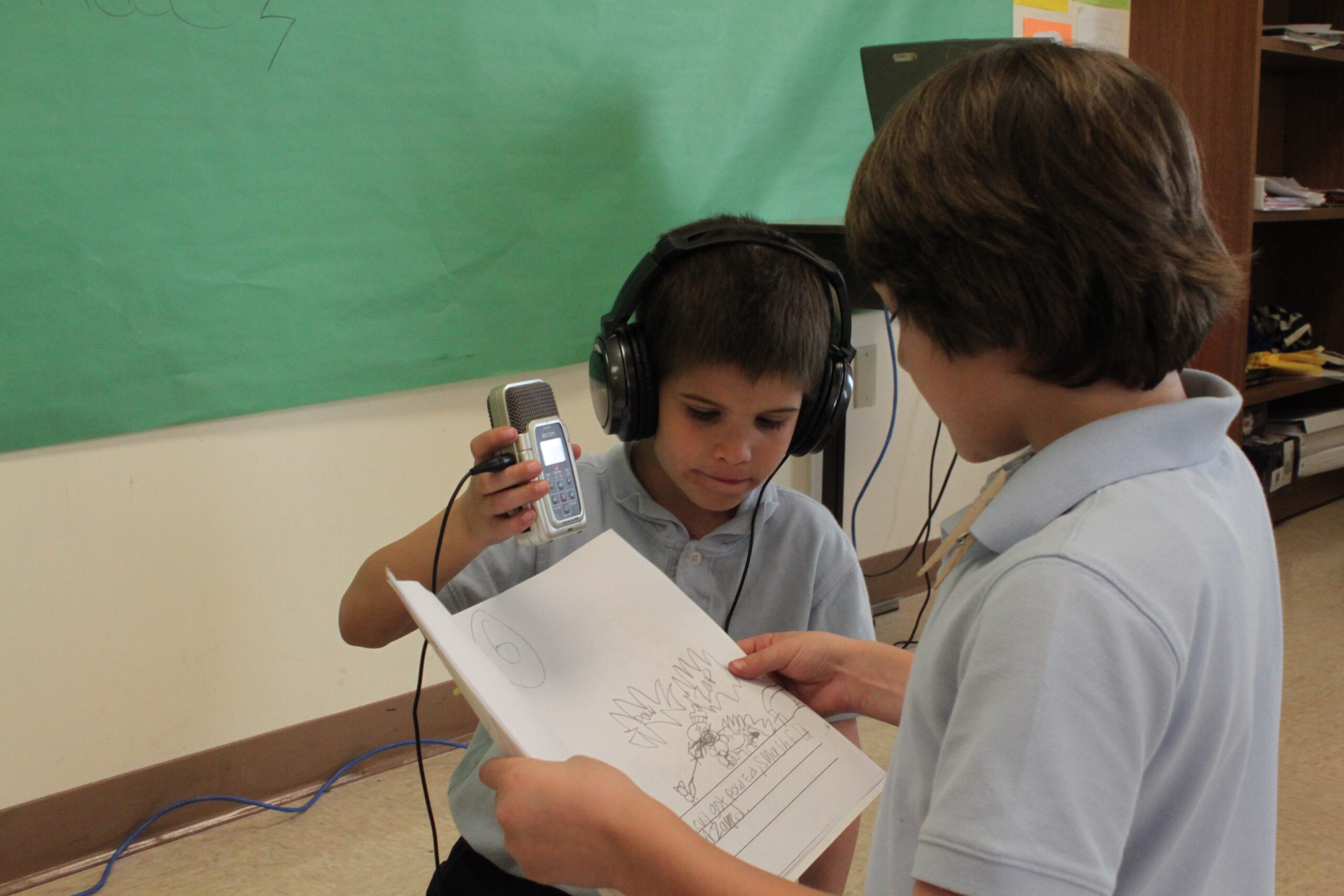 A youth wearing headphones holding a portable recorder and a youth looking at a piece of paper in a classroom
