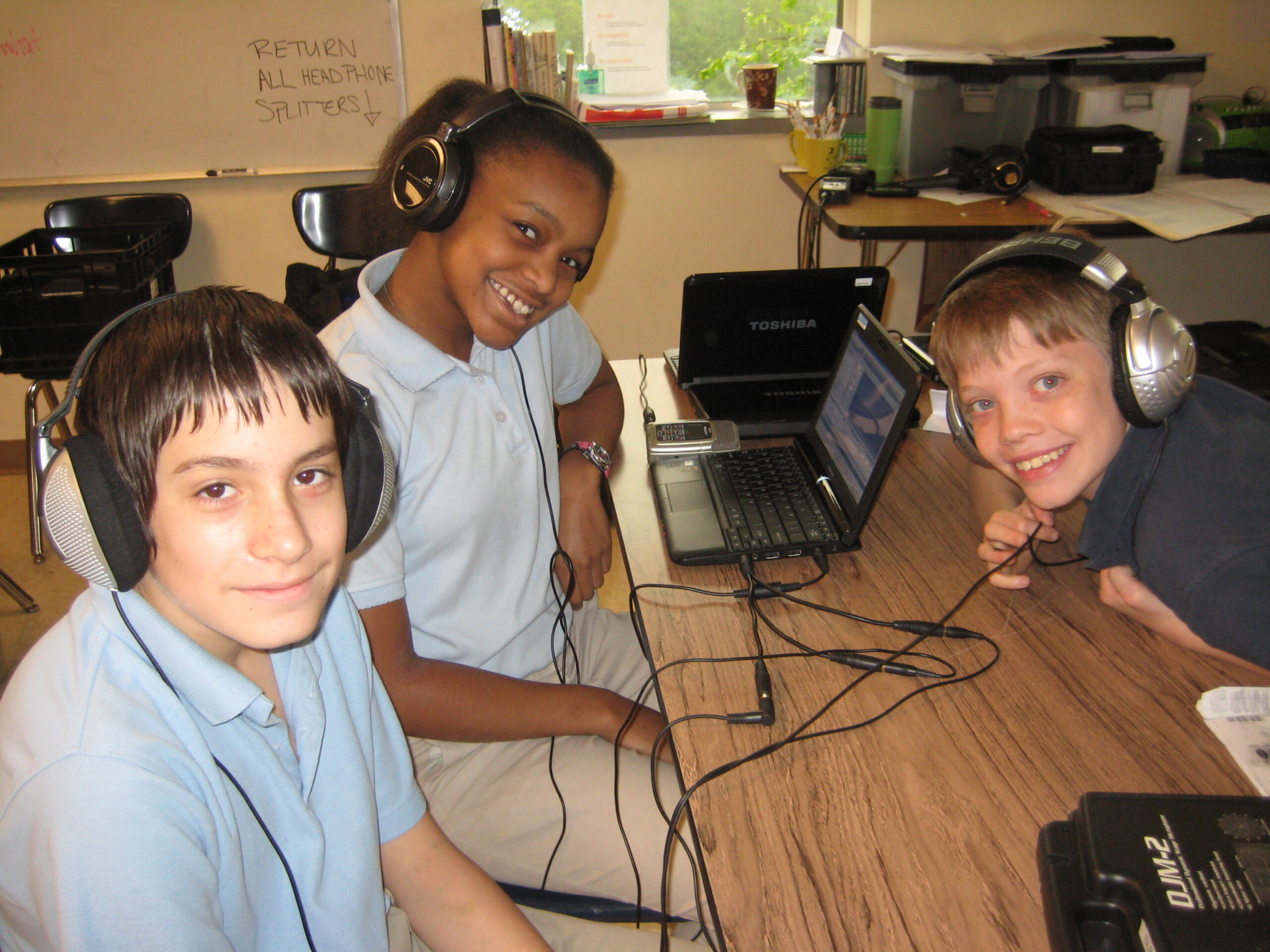 Three youth wearing headphones sitting around a laptop computer and smiling at the camera