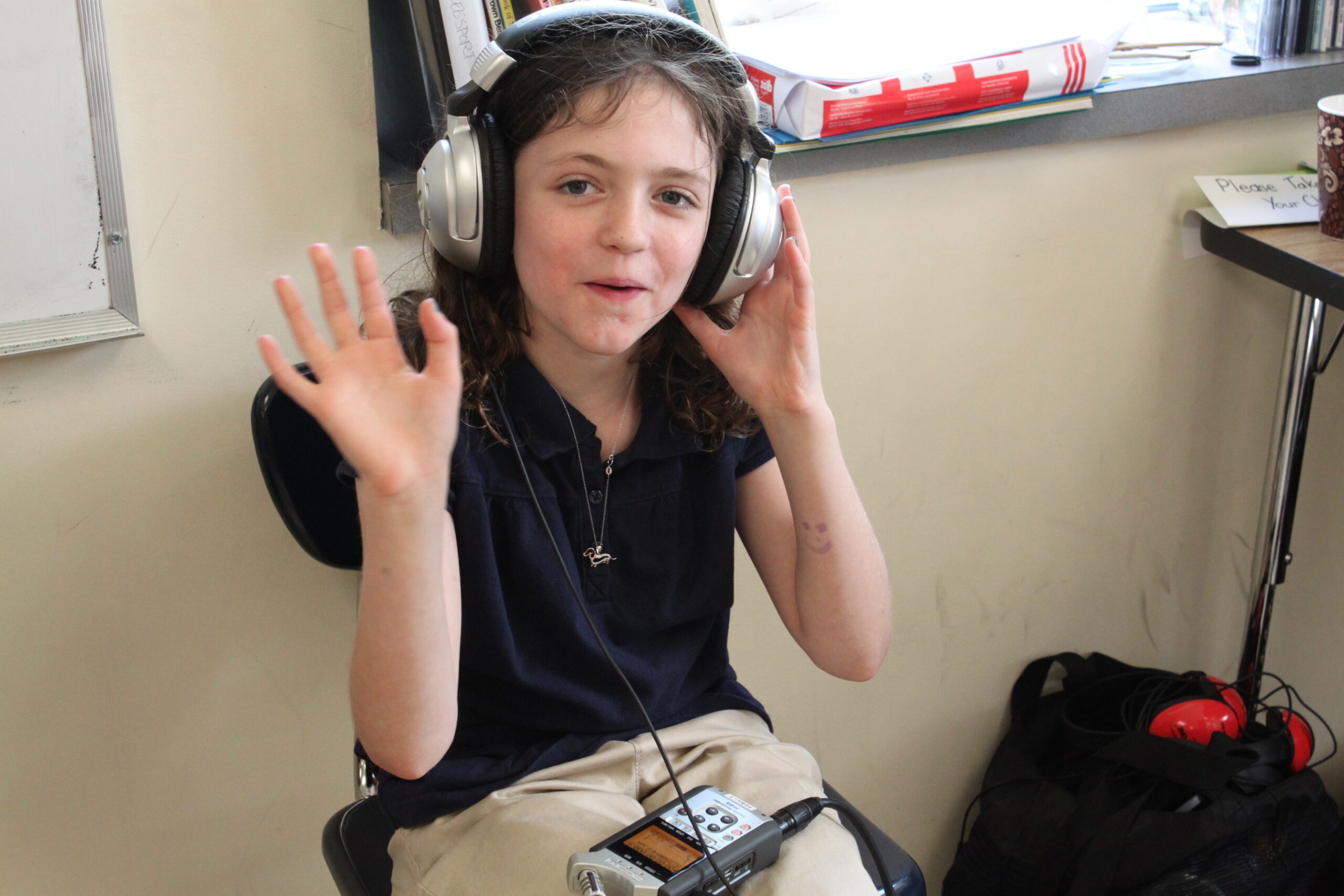 A youth wearing headphones connected to a portable recorder on their lap waving at the camera