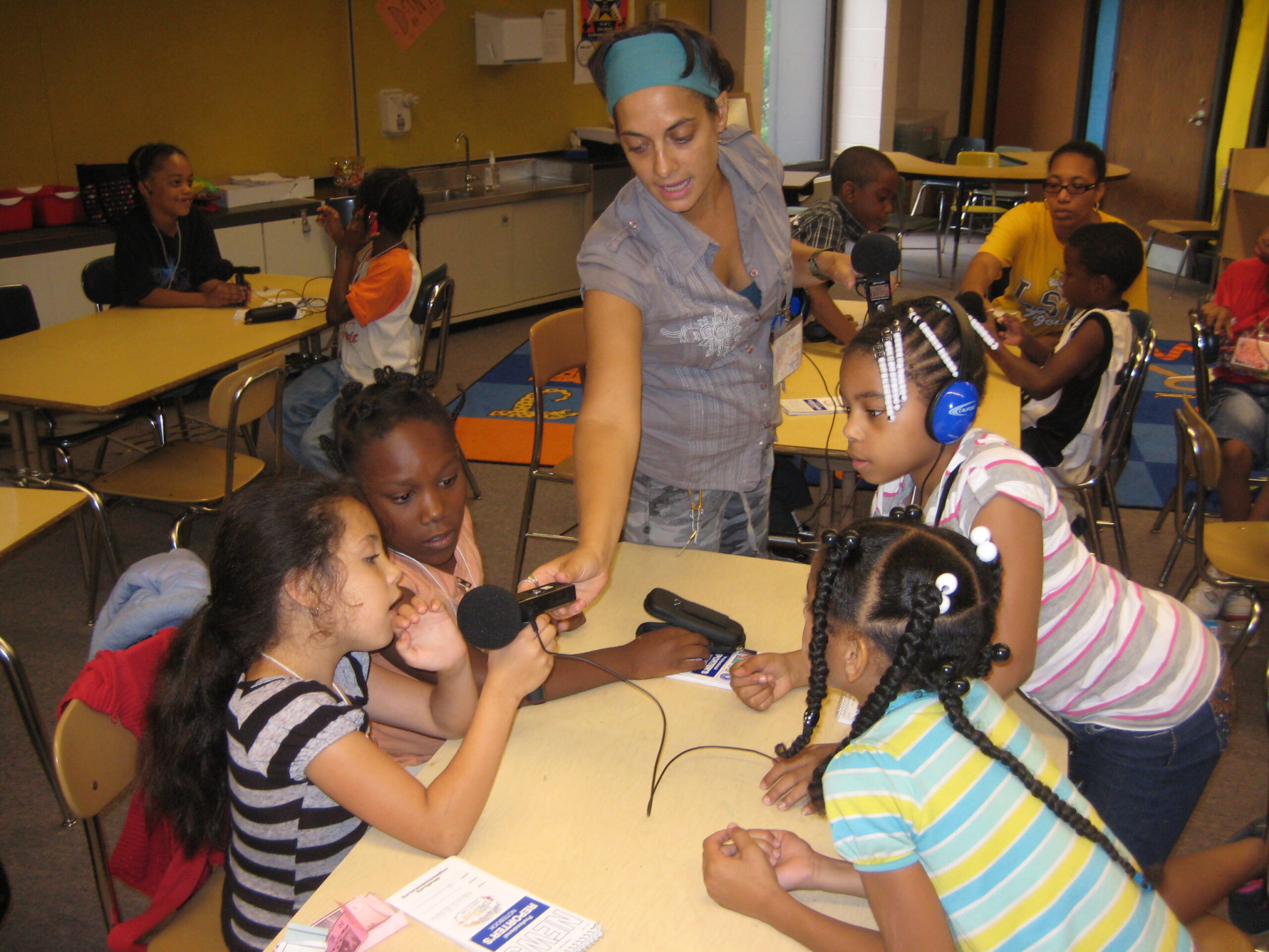 An adult holding a microphone for a youth to speak into and three other youth sitting around a table, one wearing blue headphones