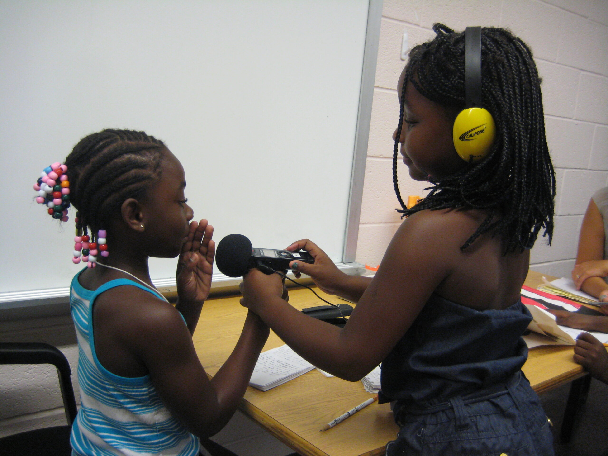 A youth wearing yellow headphones holding a portable recorder up to another youth speaking in a classroom
