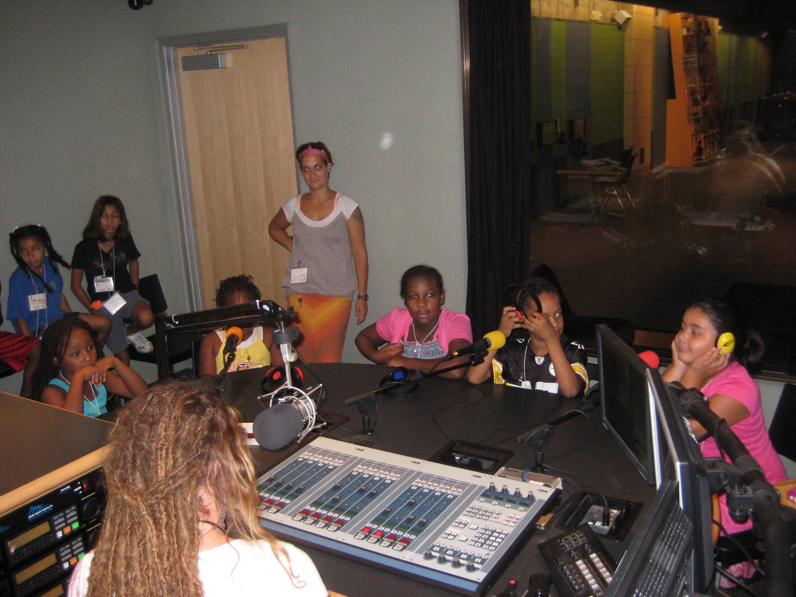 Seven youth and an adult around a table behind microphones and an adult sitting behind a microphone and an audio mixer in a radio studio