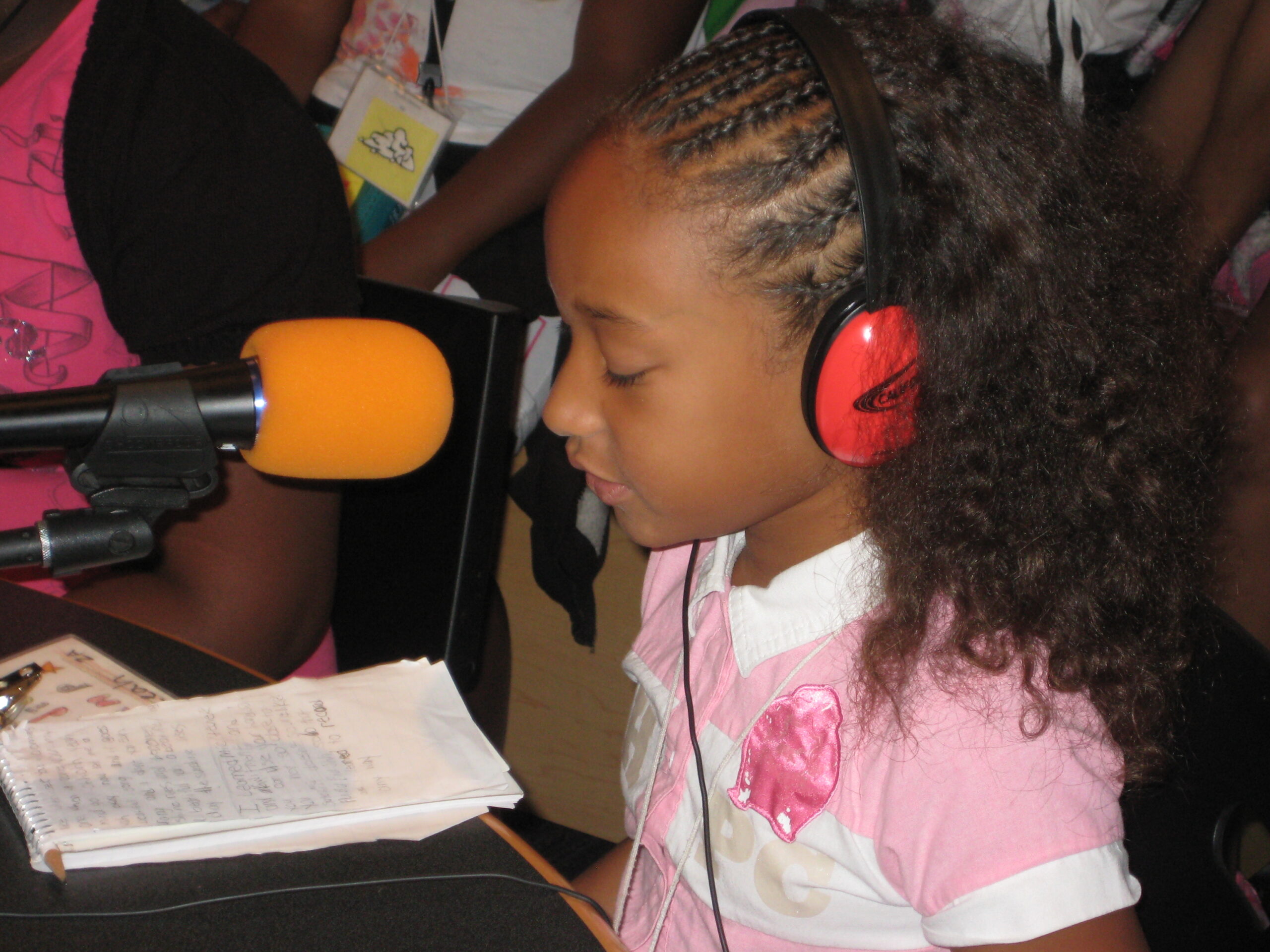 A youth wearing red headphones and speaking into a microphone while reading a piece of paper