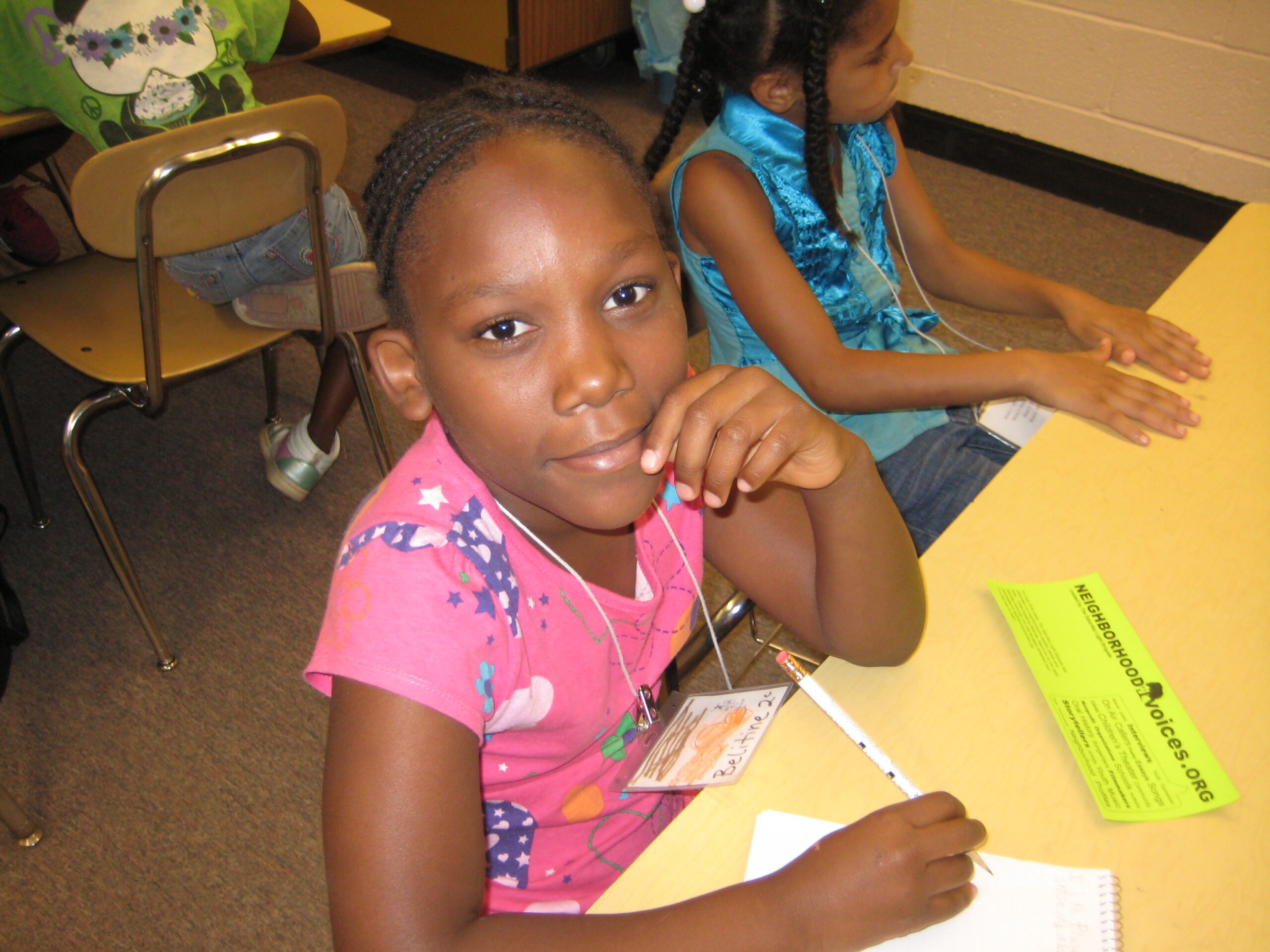 A youth looking at the camera while sitting and writing on a piece of paper in a classroom