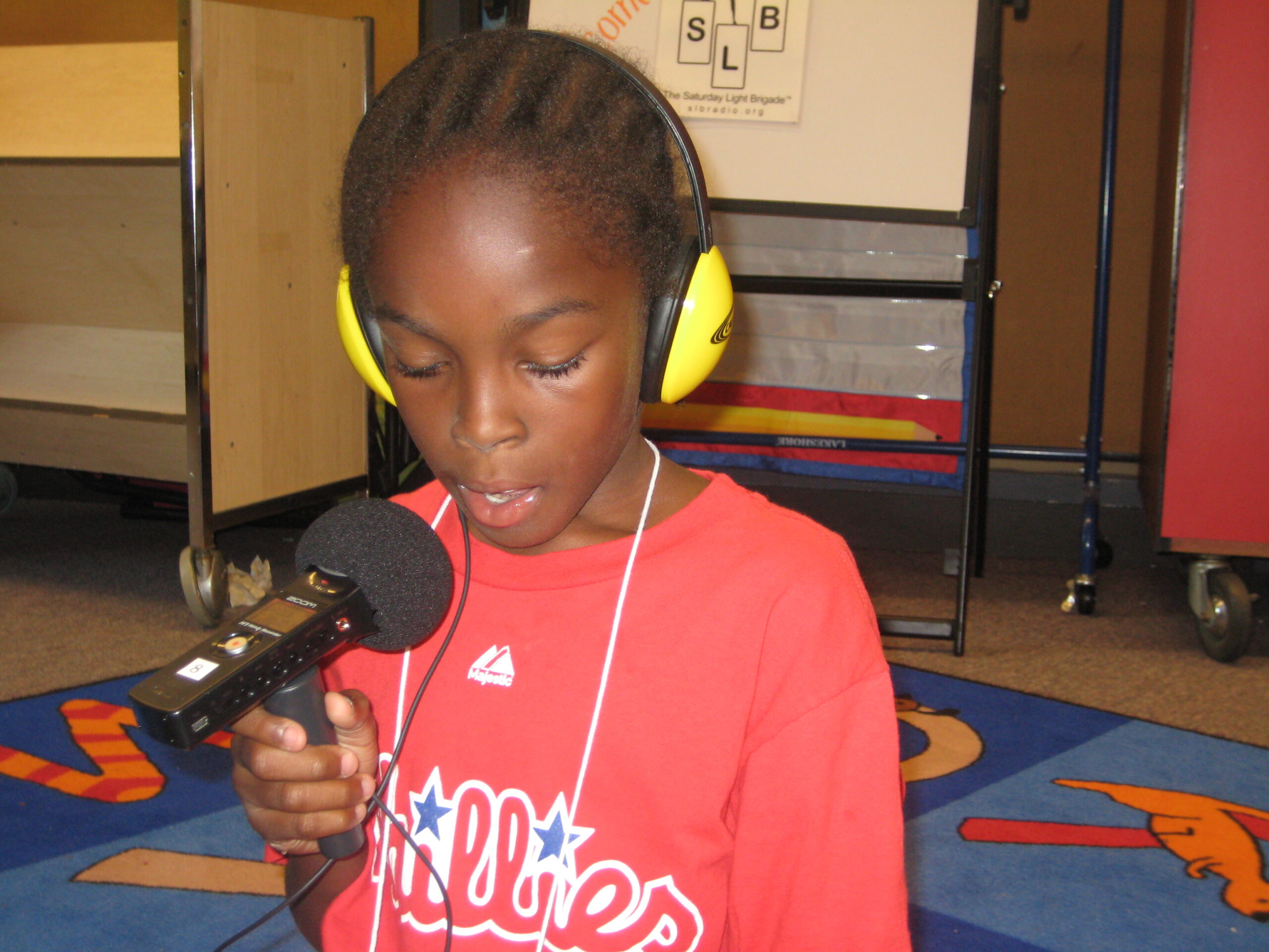 A youth wearing yellow headphones and speaking into a portable recorder in a classroom
