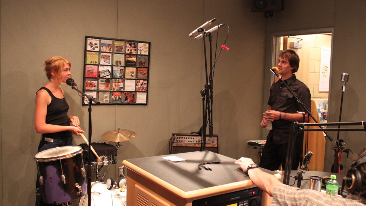 An adult standing behind a drum set and an adult singing into a microphone in a radio studio