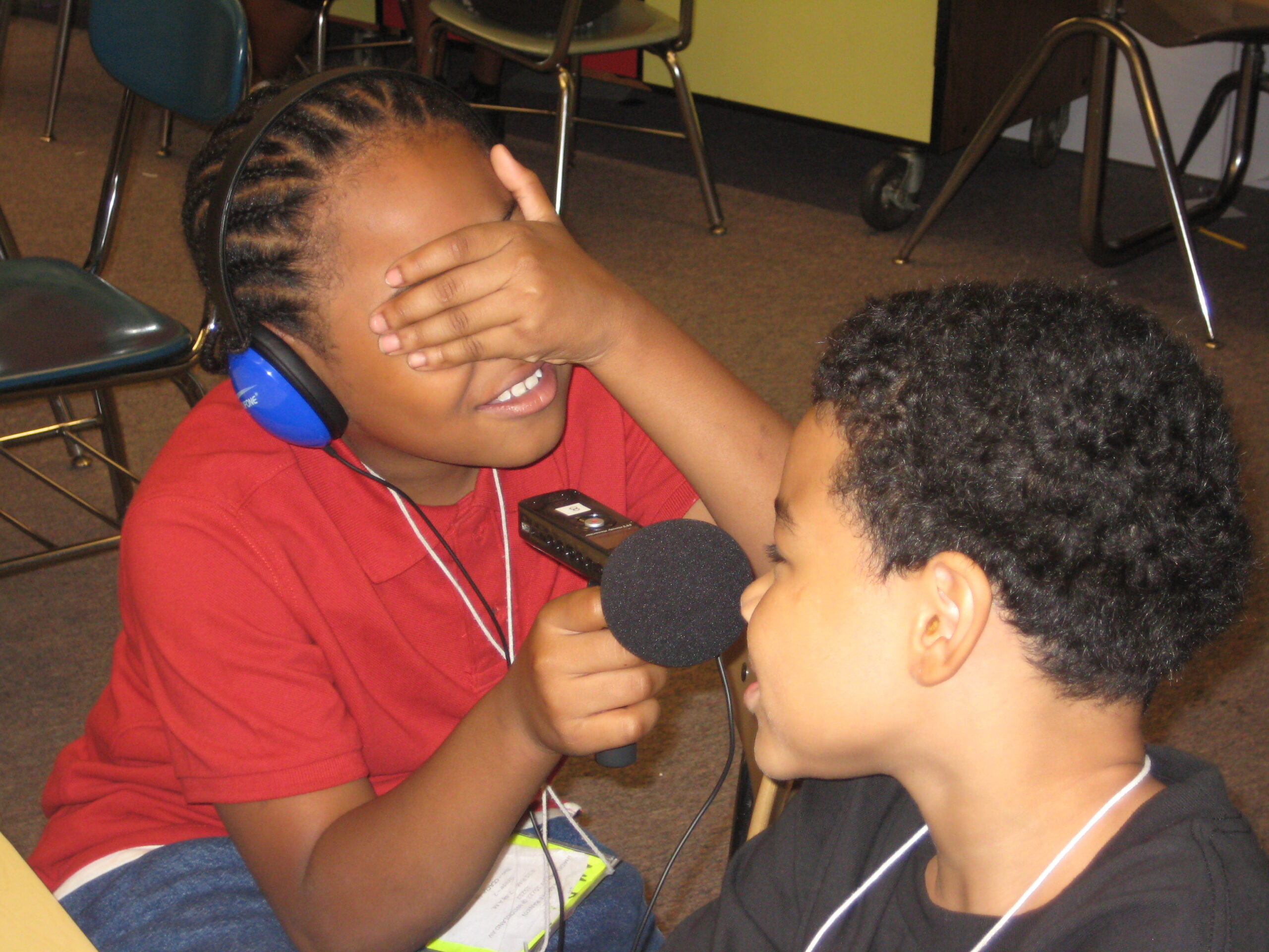 A youth wearing headphones holding their face in their hand and holding a microphone up to another youth