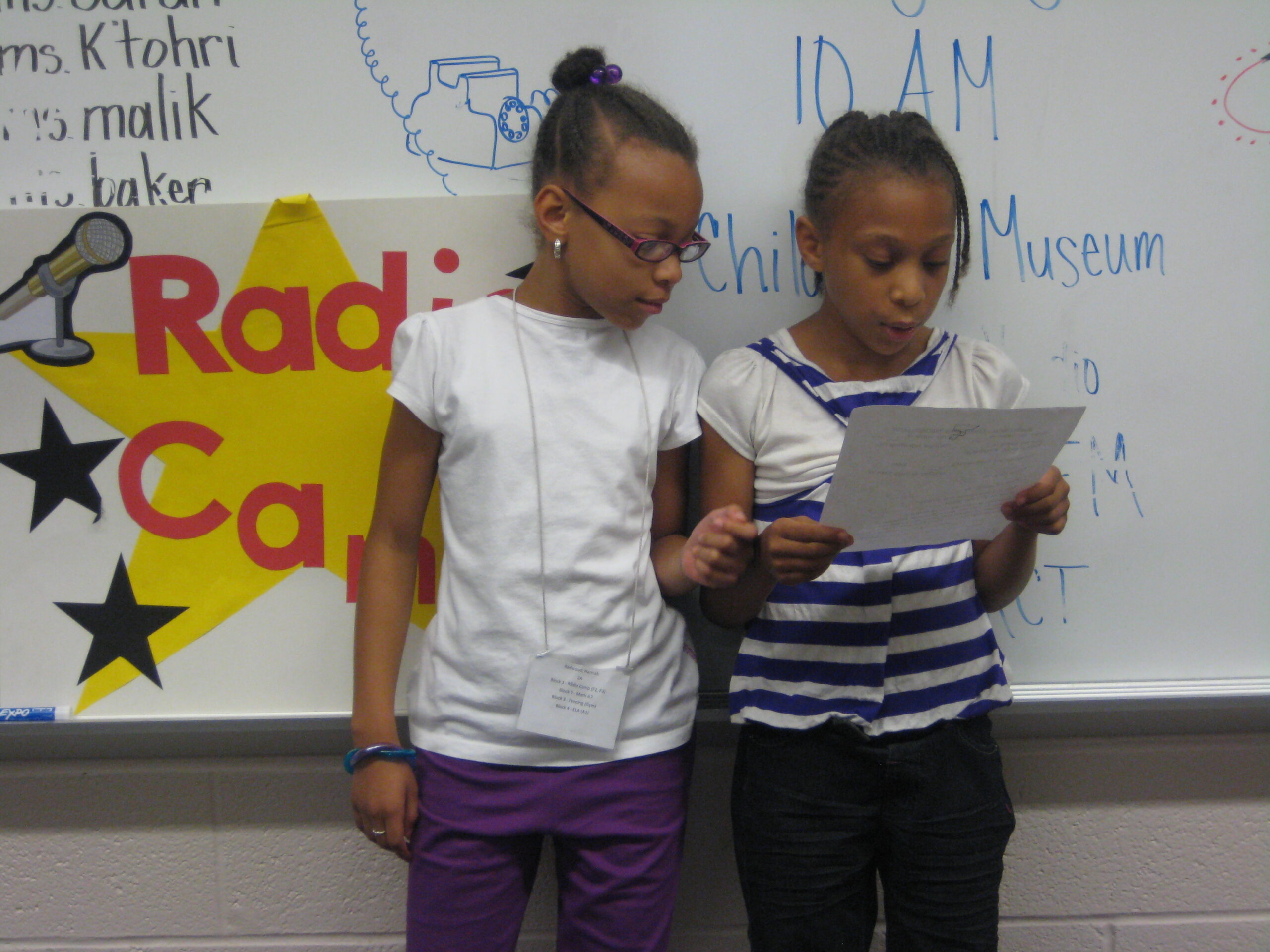Two youth looking at a piece of paper while standing in front of a white board in a classroom