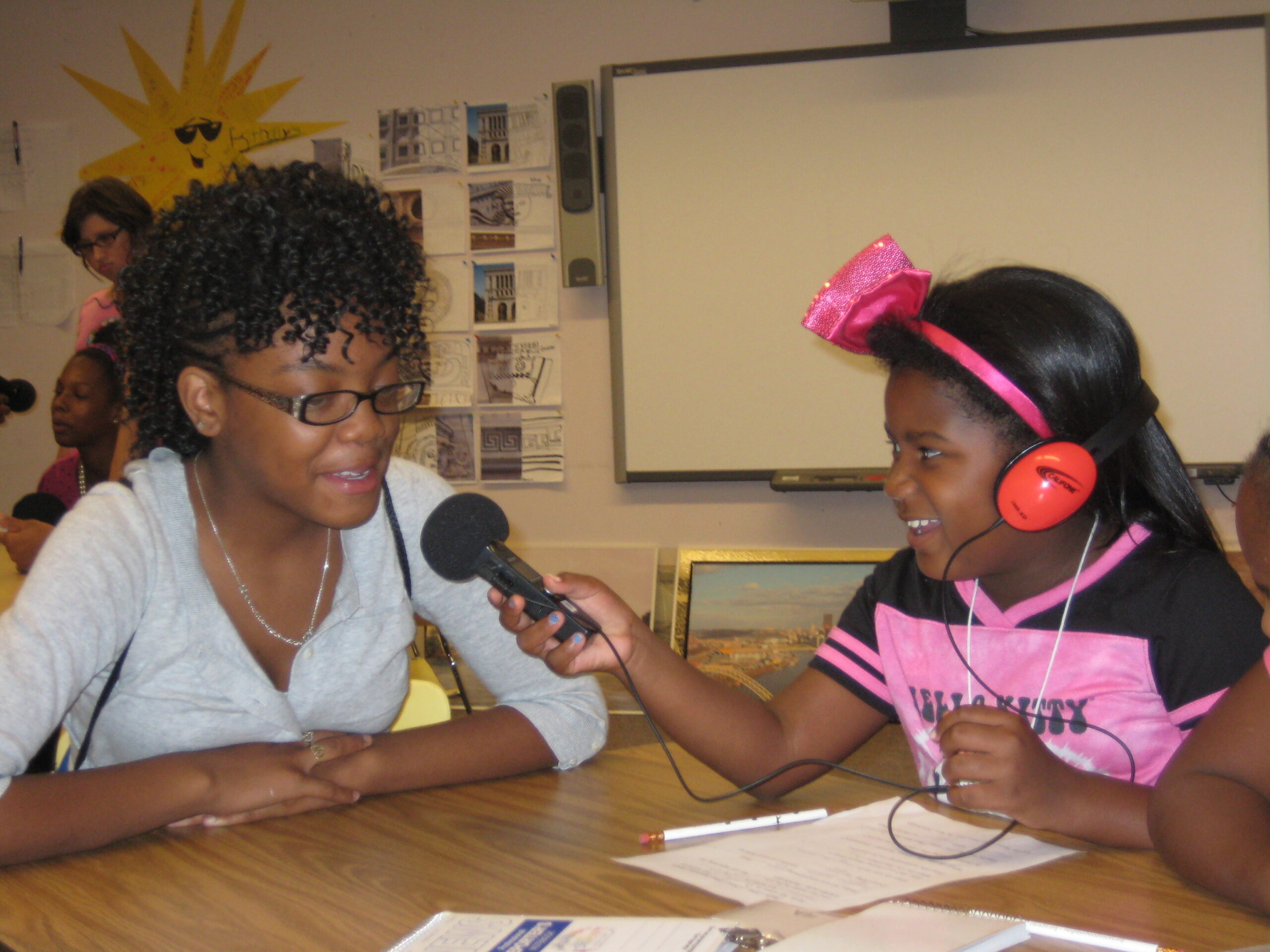 A youth wearing headphones and holding a microphone interviewing an adult while sitting at a table in a classroom
