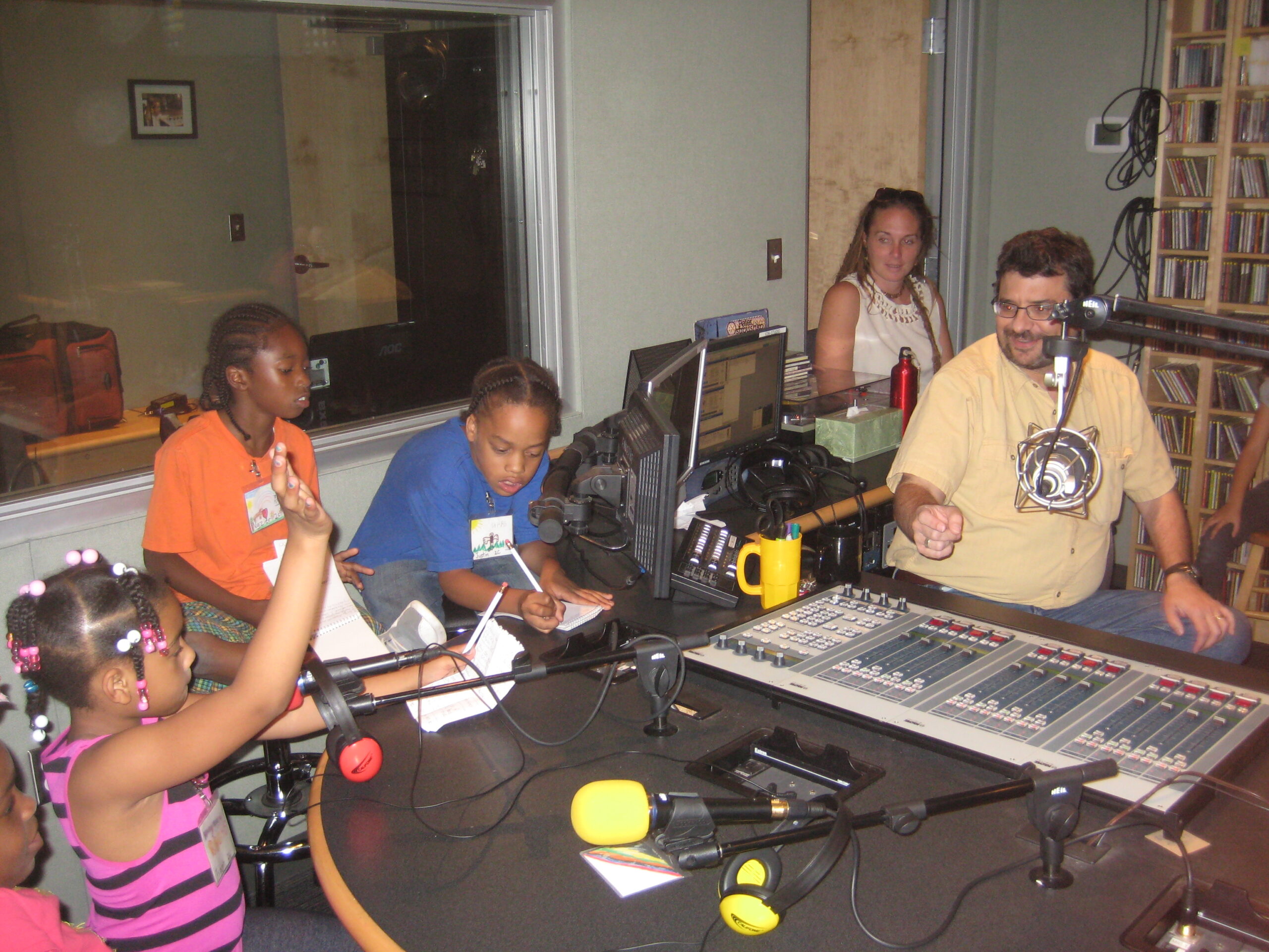 Three youth sitting behind microphones and two adults sitting behind a microphone and an audio mixer in a radio studio