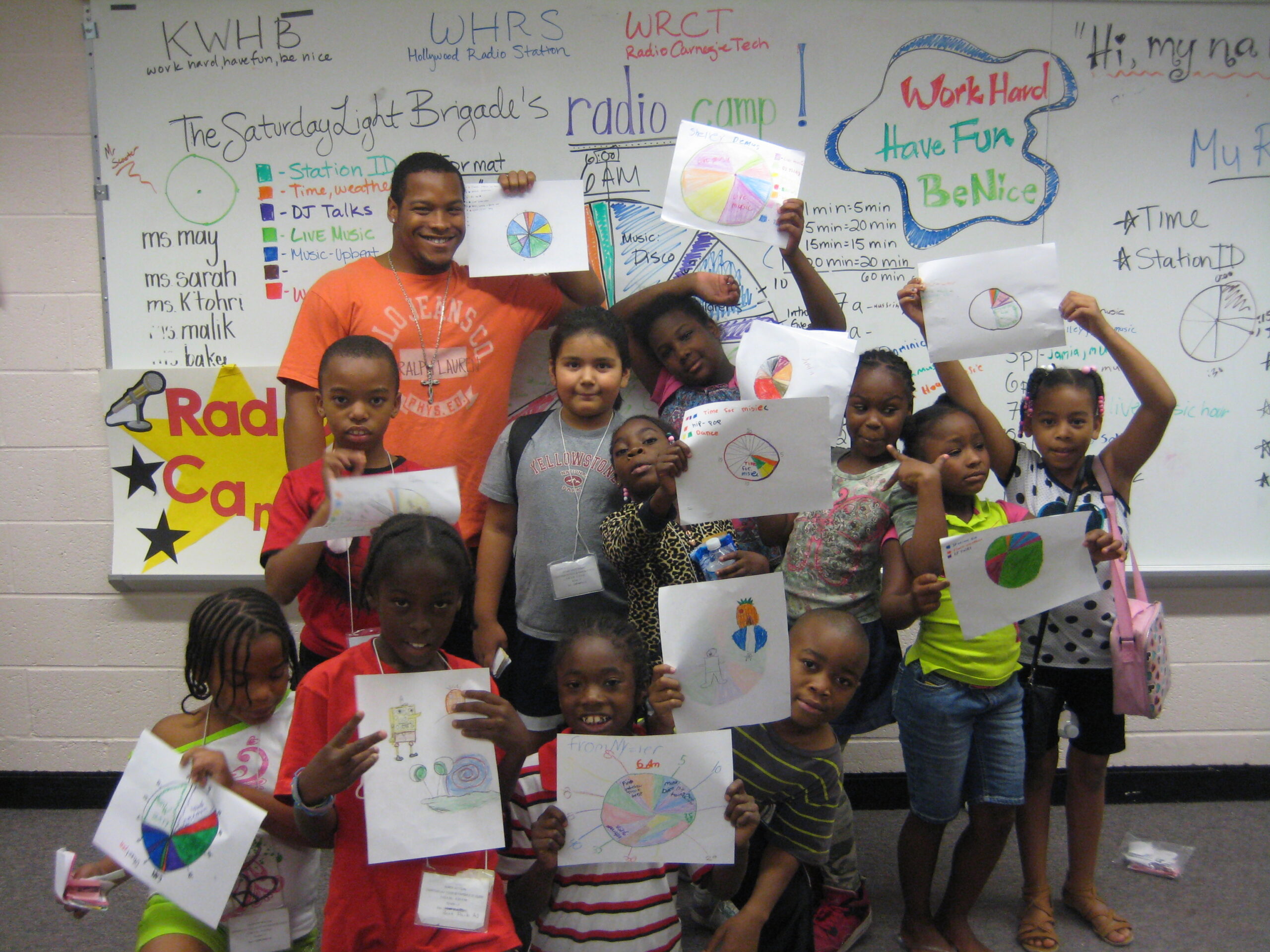 Ten youth and an adult holding up pieces of paper in front of a whiteboard in a classroom