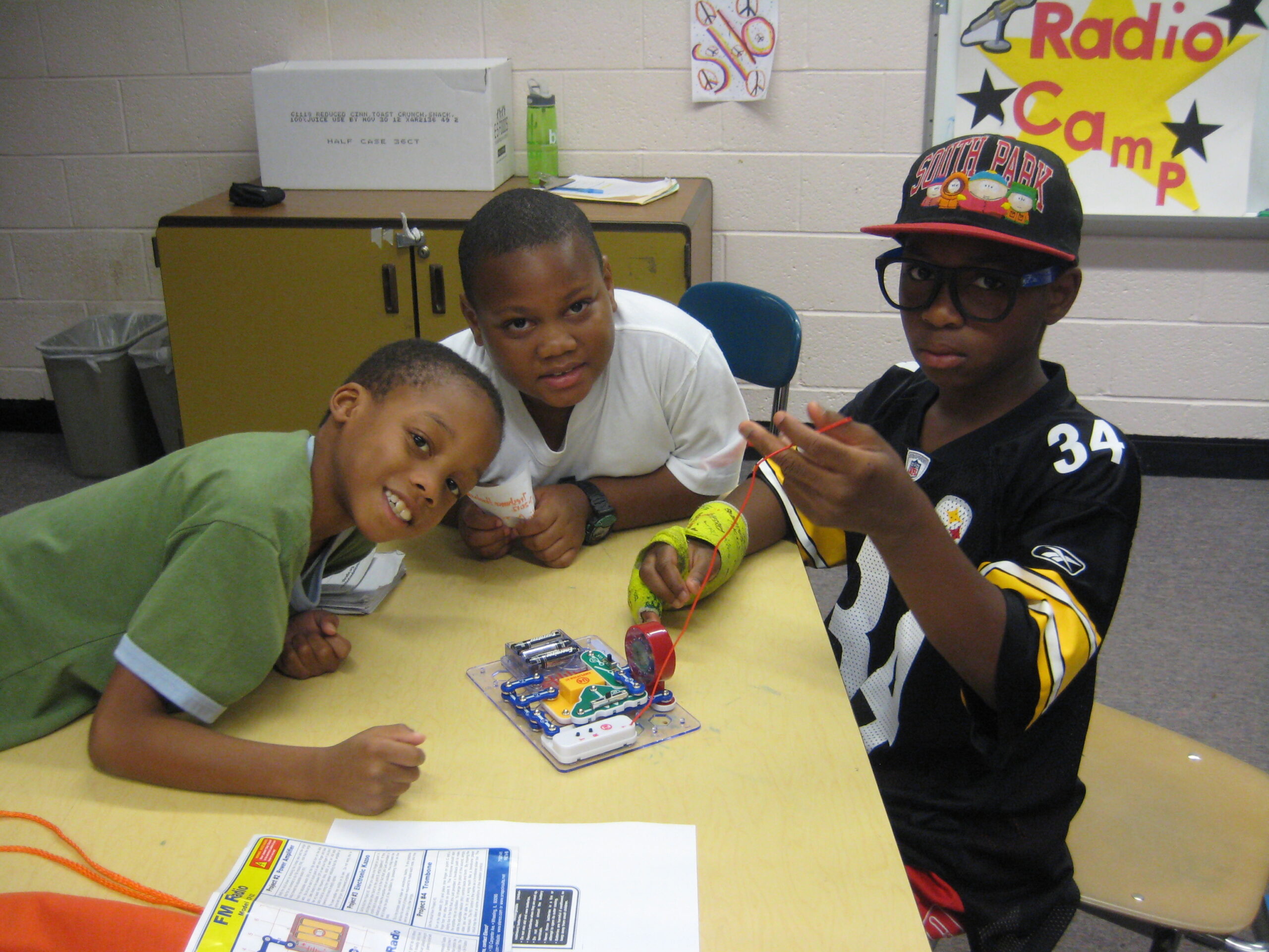 Three youth sitting around an electrical contraption on a classroom table