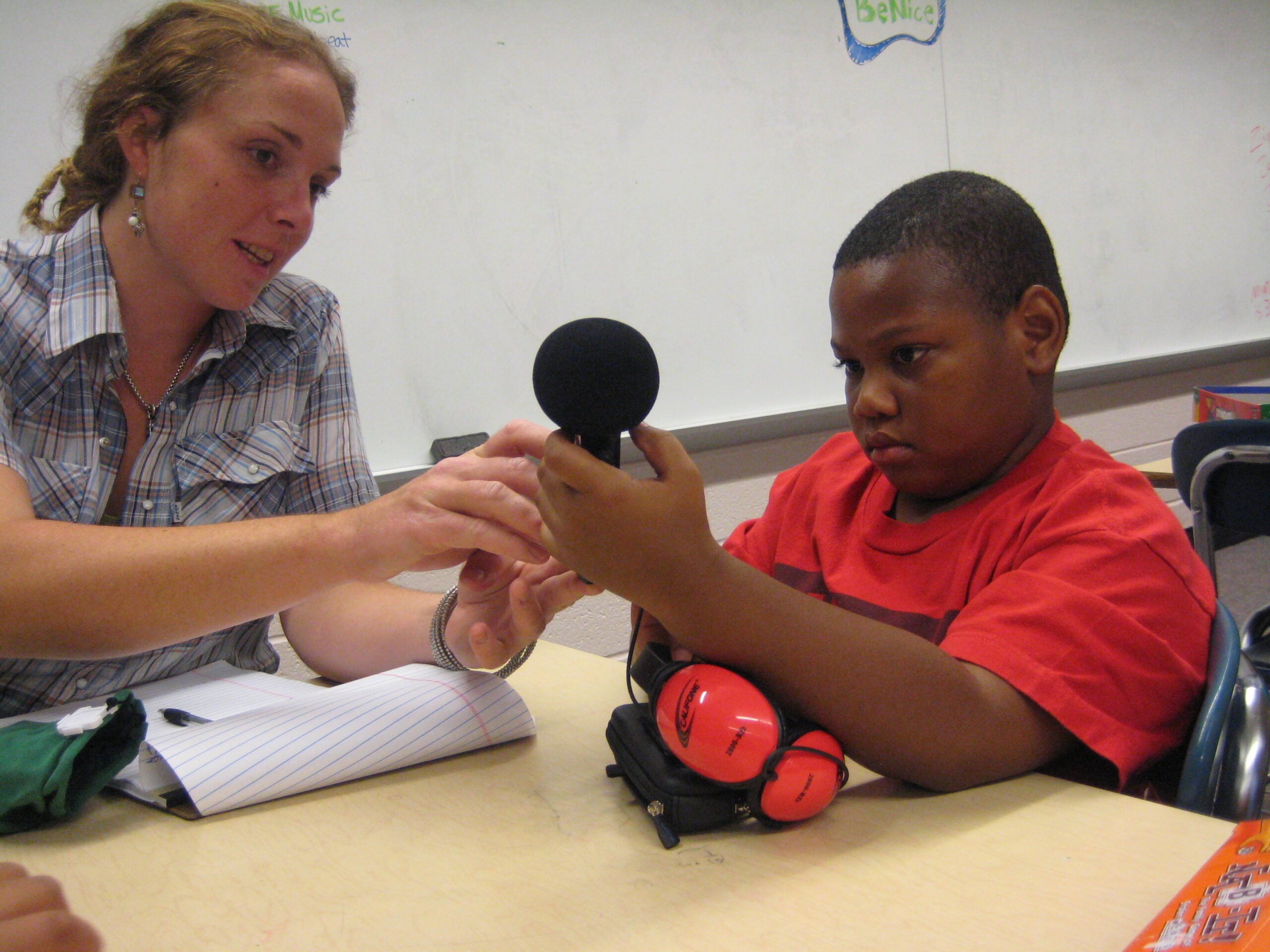 An adult and a child holding a microphone together while sitting in front of a whiteboard in a classroom