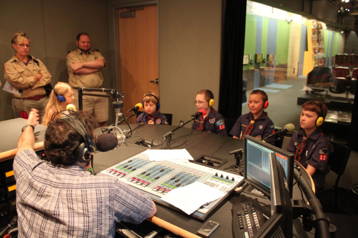 Four youth in uniform wearing headphones speaking into microphones being interviewed by an adult wearing headphones sitting behind a microphone and a mixing board as two adults in uniform look on in a radio studio