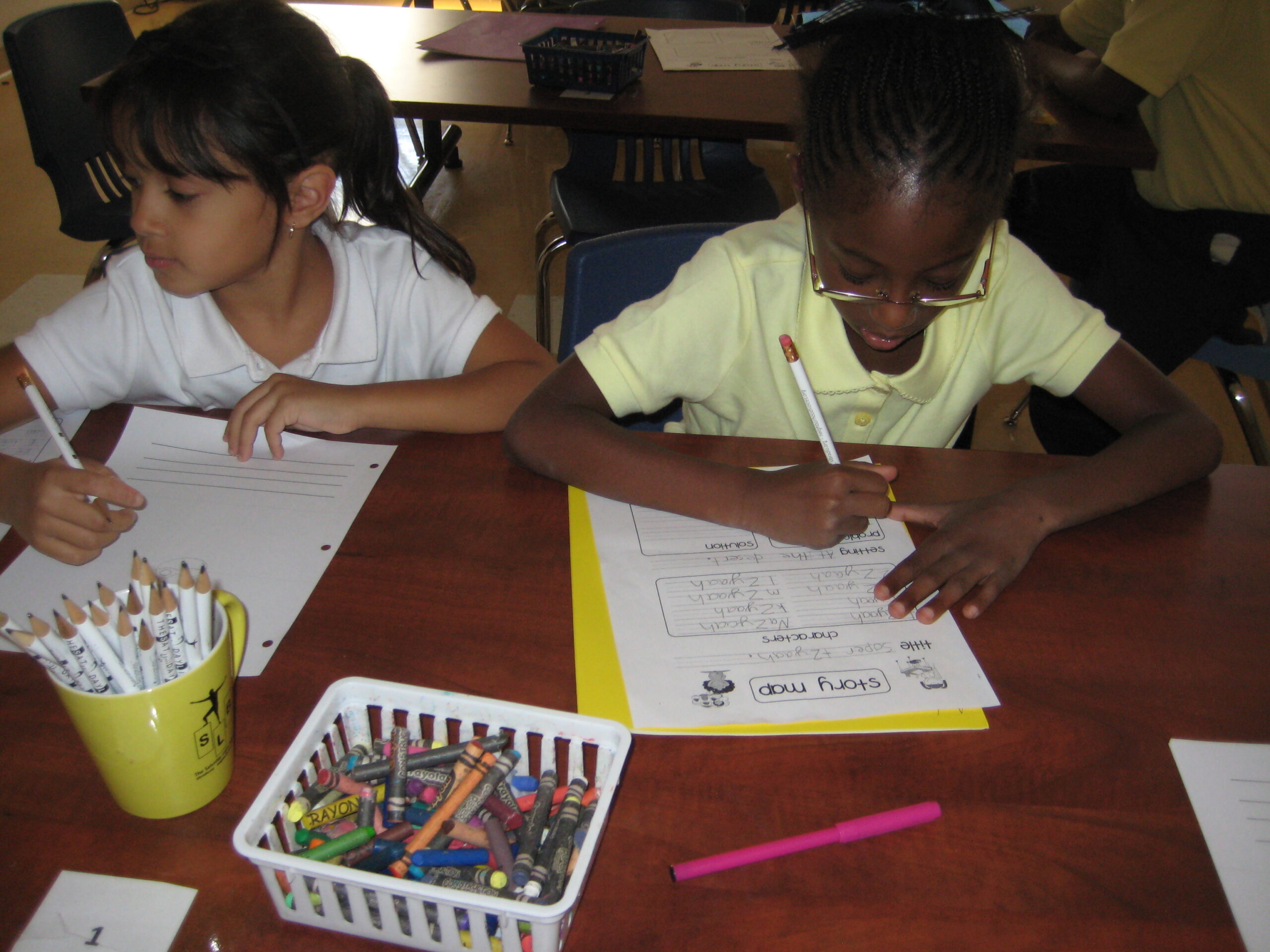Two youth writing on pieces of paper while sitting at a classroom table