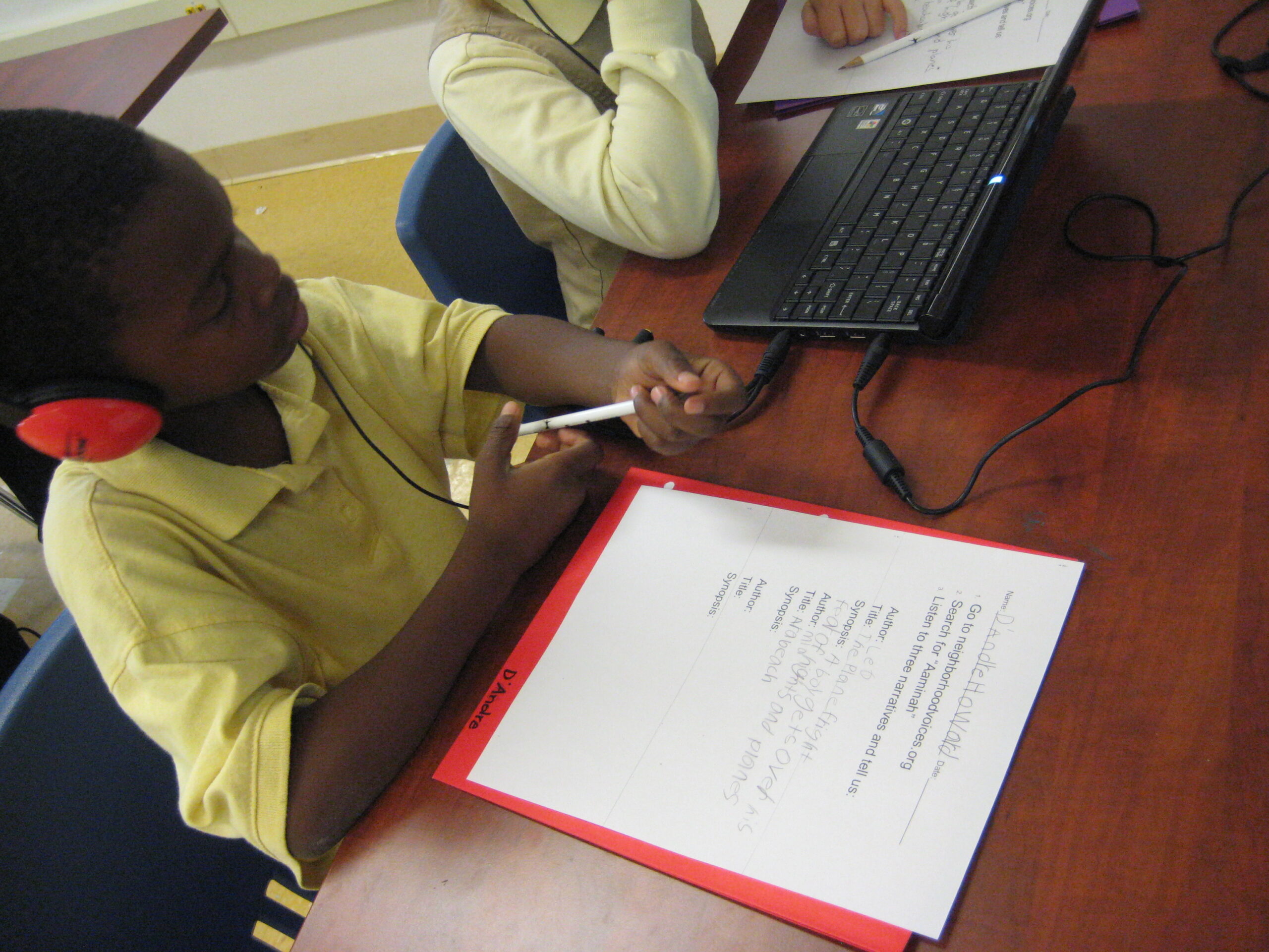 A youth wearing headphones looking at a laptop computer while sitting behind a piece of paper on a classroom table