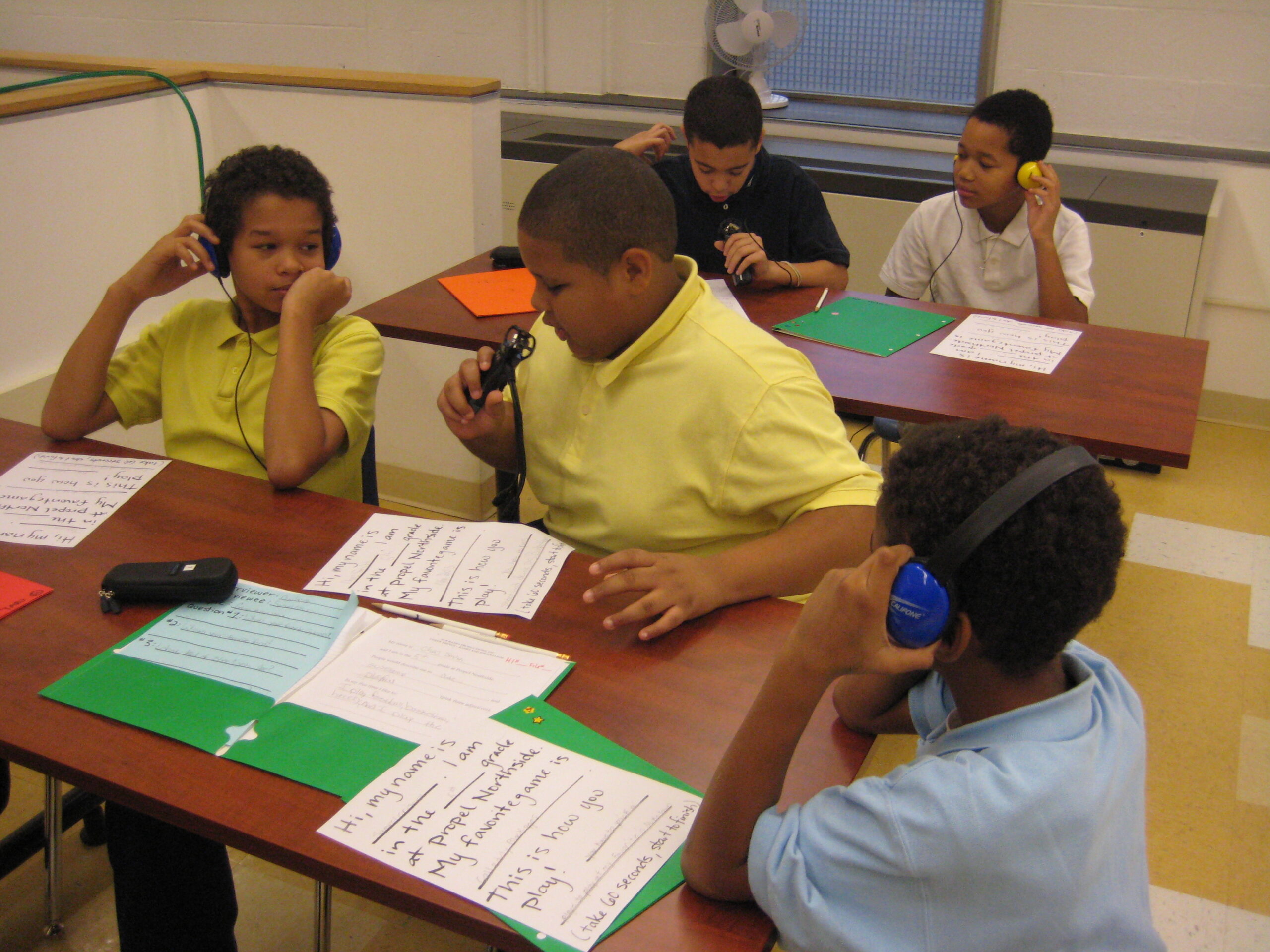 Three youth wearing headphones and two youth speaking into microphones while looking down at pieces of paper at classroom tables