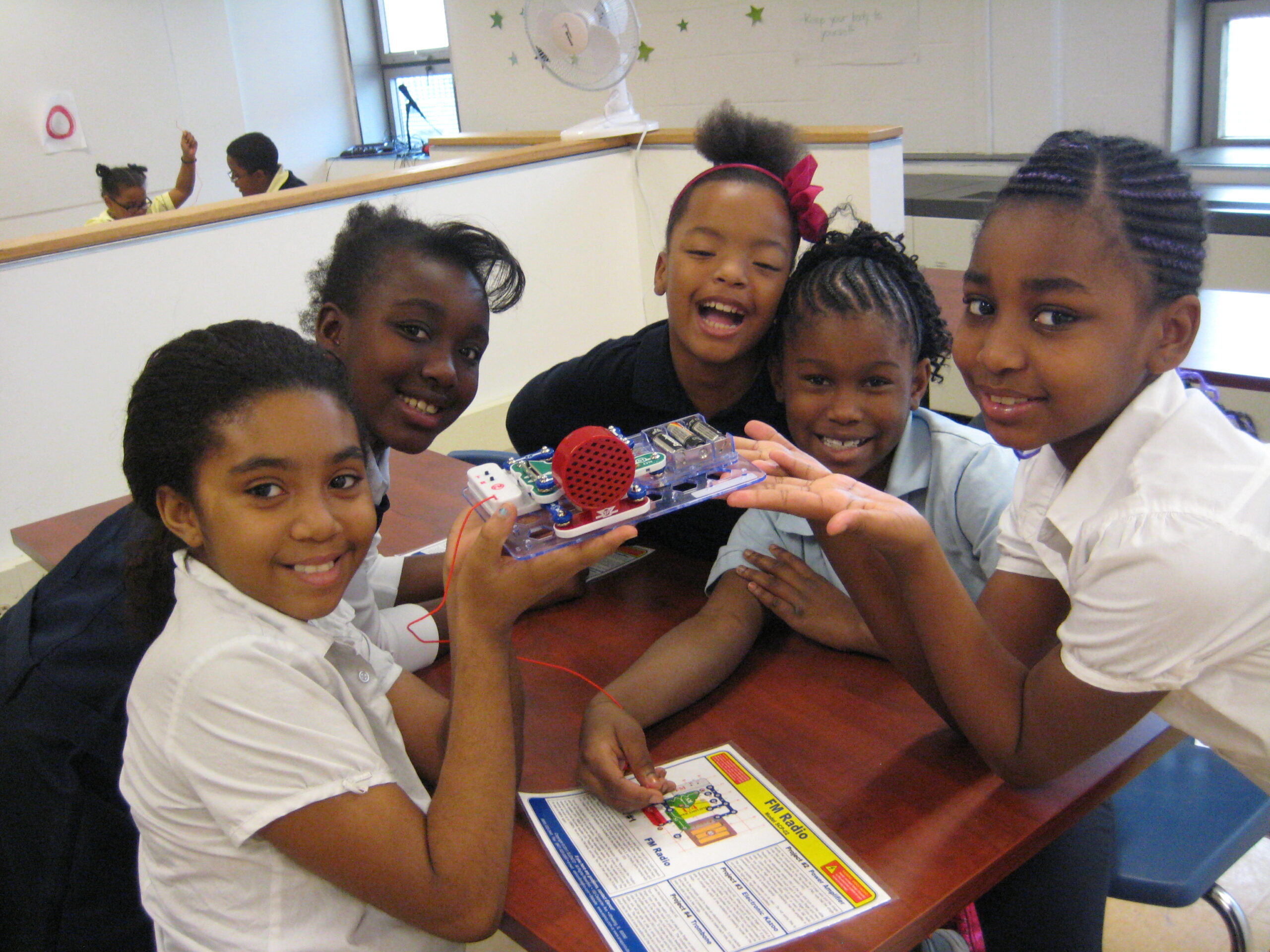 Five youth smiling and holding an electrical contraption in a classroom.