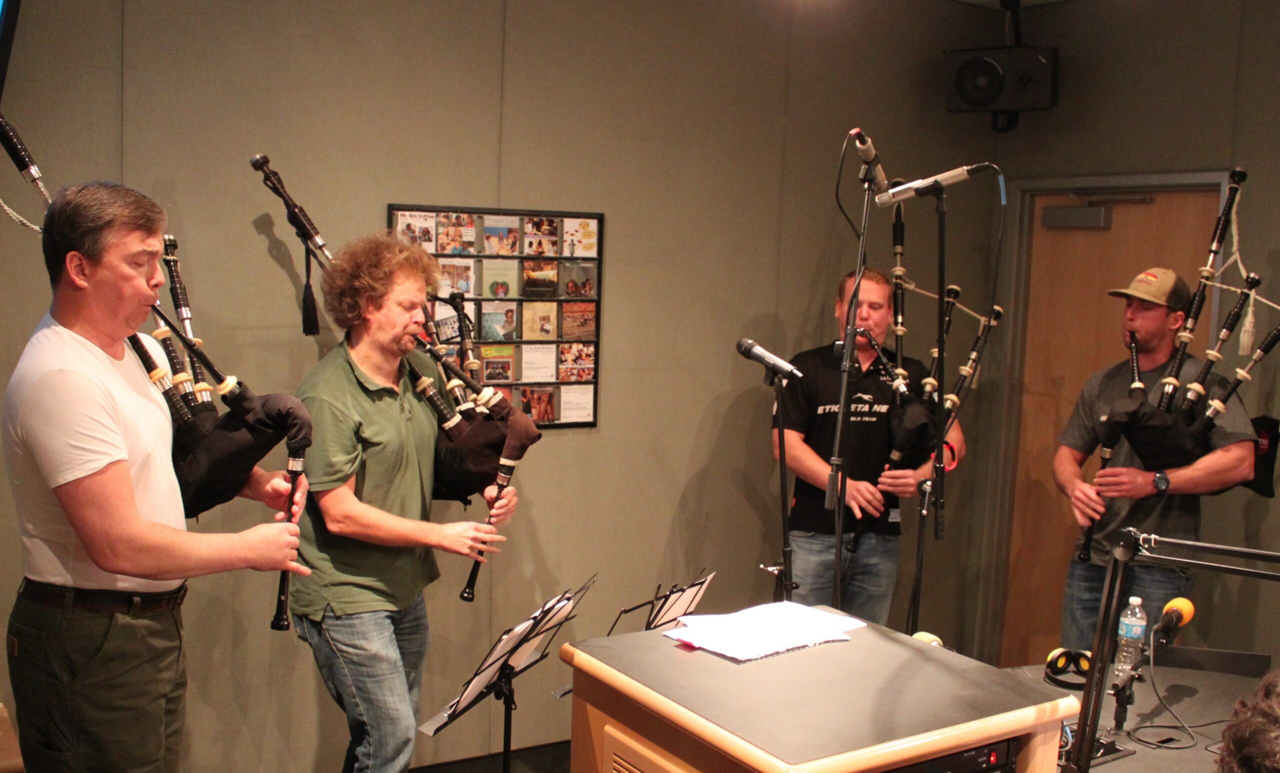 Four adults playing bagpipes while looking at sheet music on music stands in a radio studio