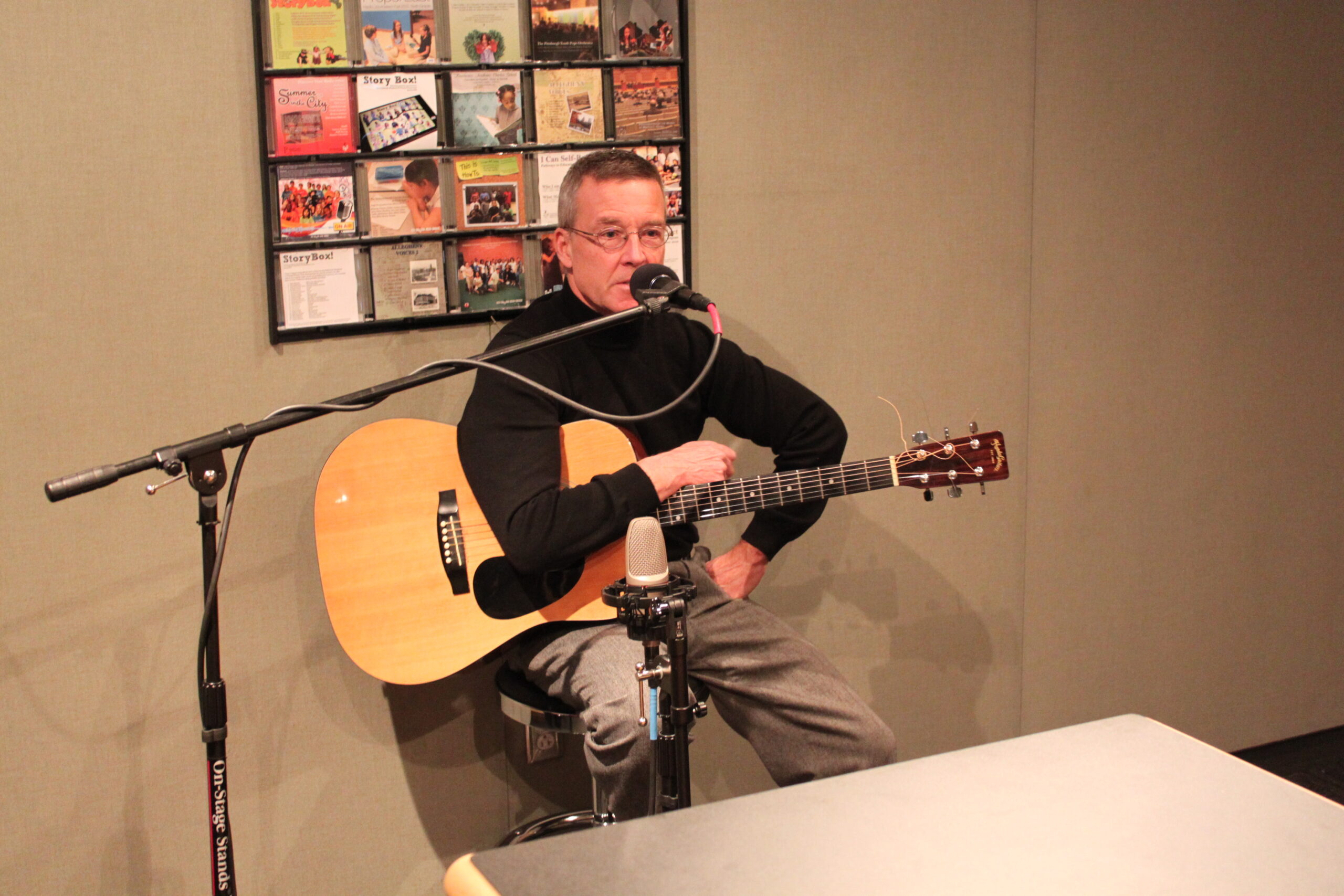 An adult sitting while holding an acoustic guitar and speaking into a microphone in a radio studio