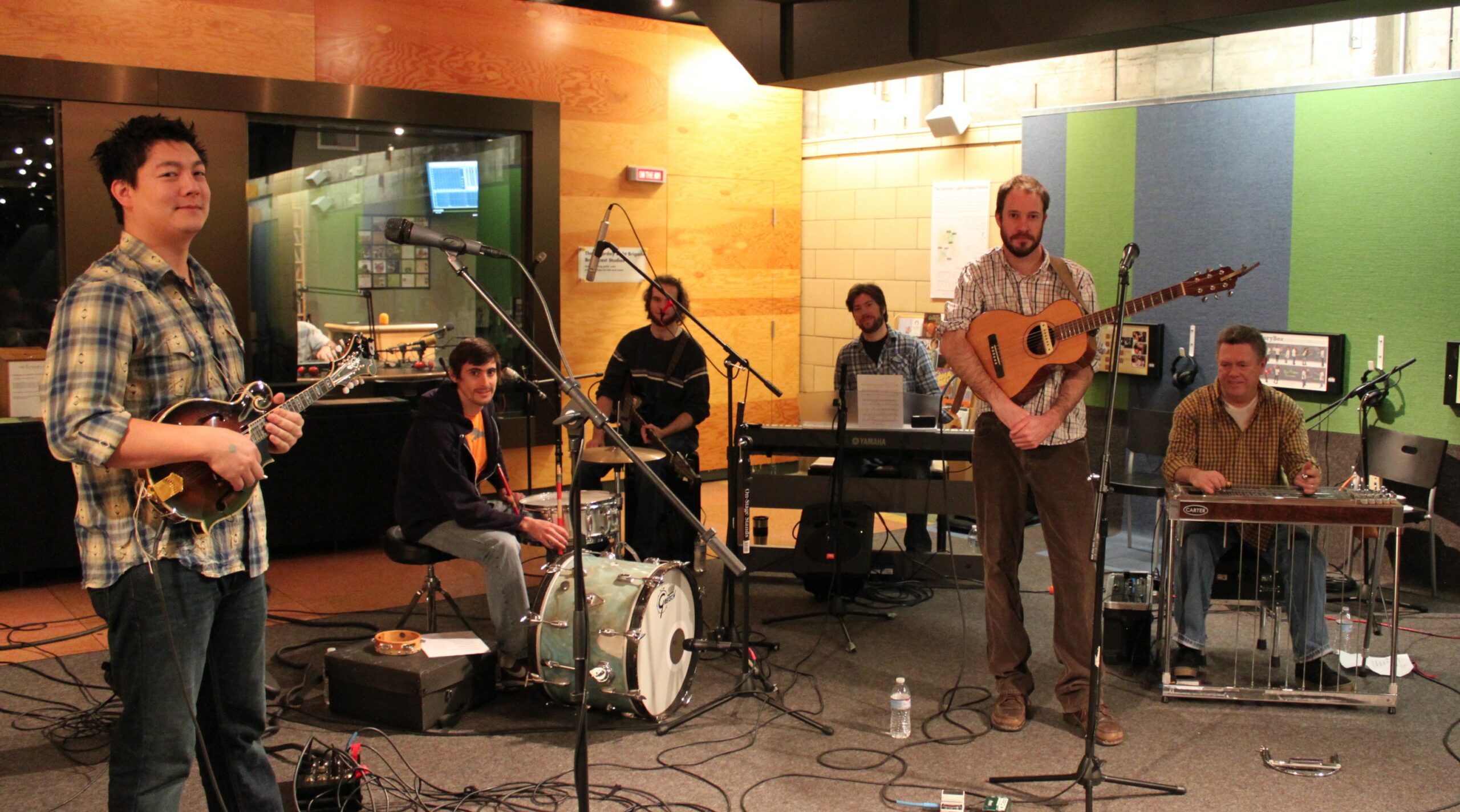 An adult holding a mandolin, an adult sitting at a drumset, an adult sitting and holding an electric bass, an adult sitting at a piano keyboard, an adult holding an acoustic guitar, and an adult sitting at a pedal steel guitar.