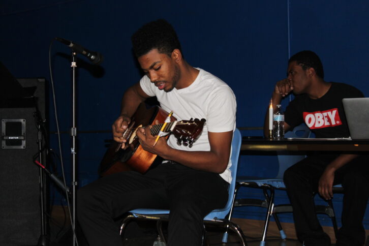 A teen playing an acoustic guitar while sitting in a chair and another teen sitting in the background listening in a blue room