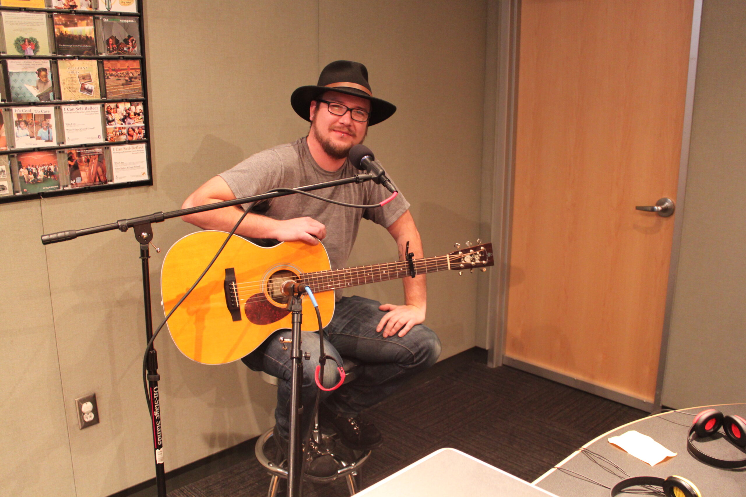 An adult sitting on a stool behind a microphone holding an acoustic guitar in a radio studio
