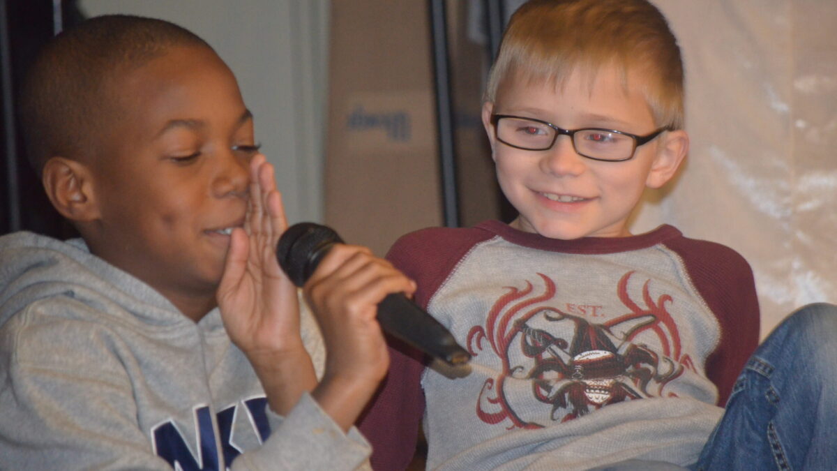 A youth smiling while looking at another youth putting their hand in between their face and a microphone