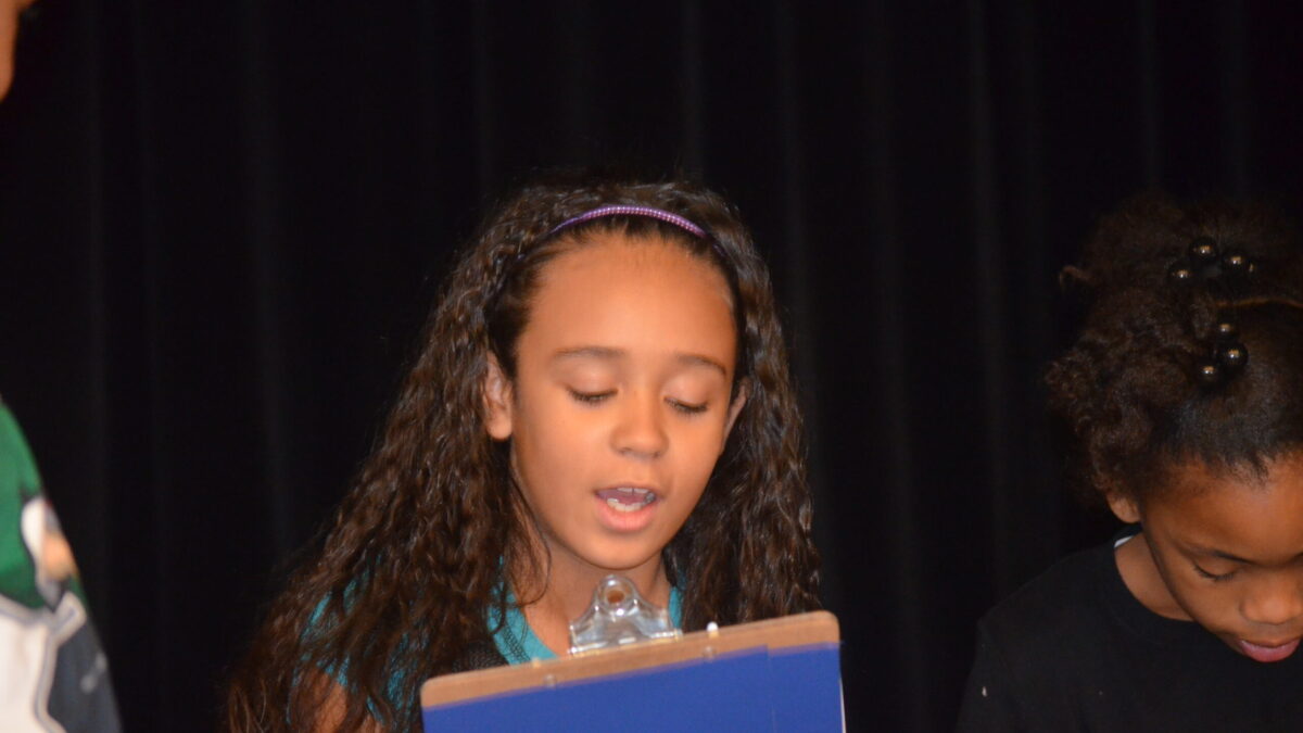 A youth reading from a blue clipboard in front of a black stage curtain