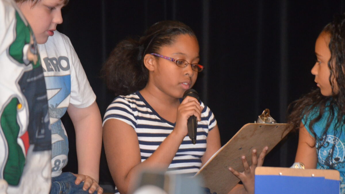 Two youth looking at another youth reading a clipboard and speaking into a held microphone