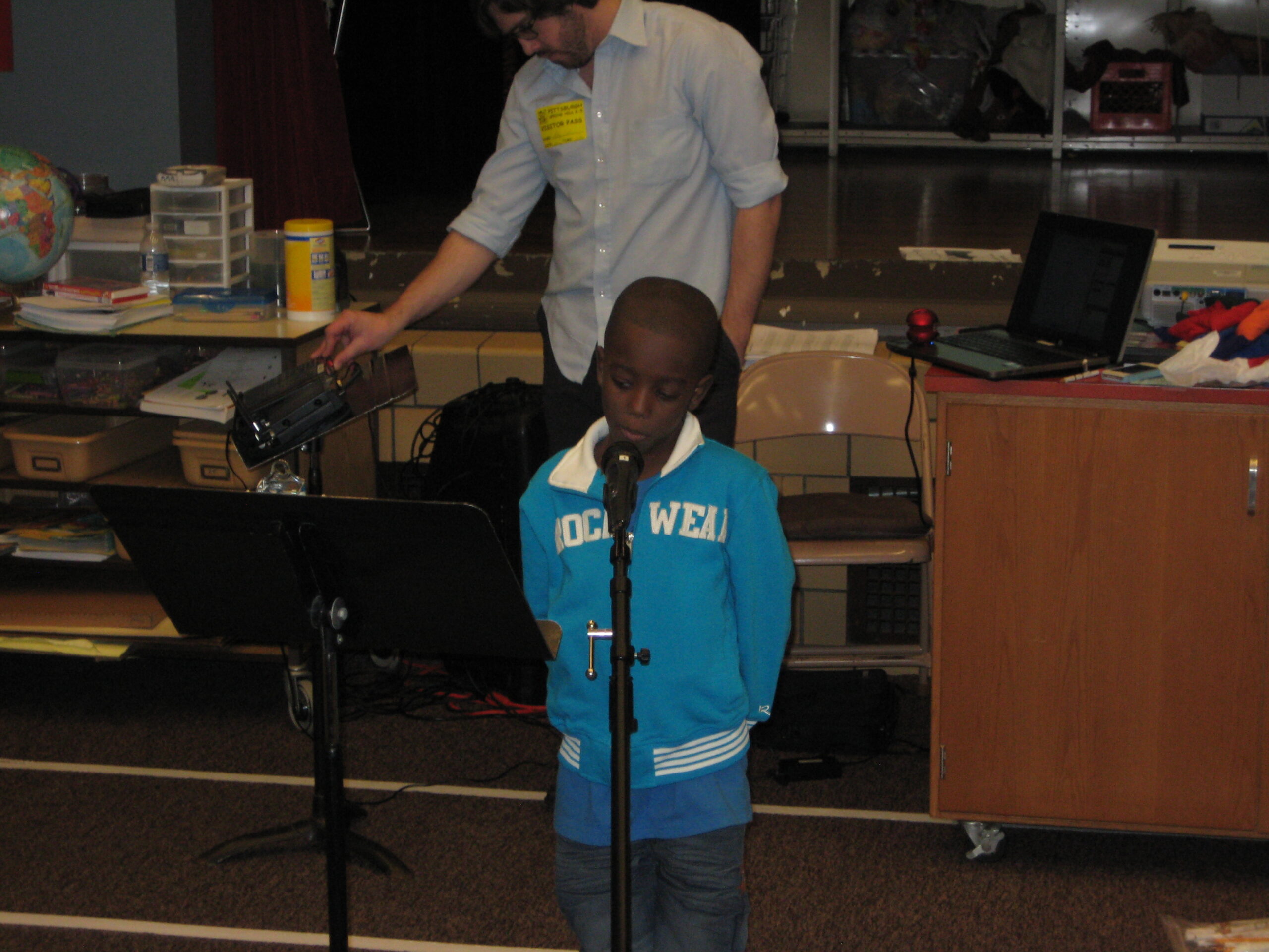 An adult standing behind a youth speaking into a microphone while looking at a music stand