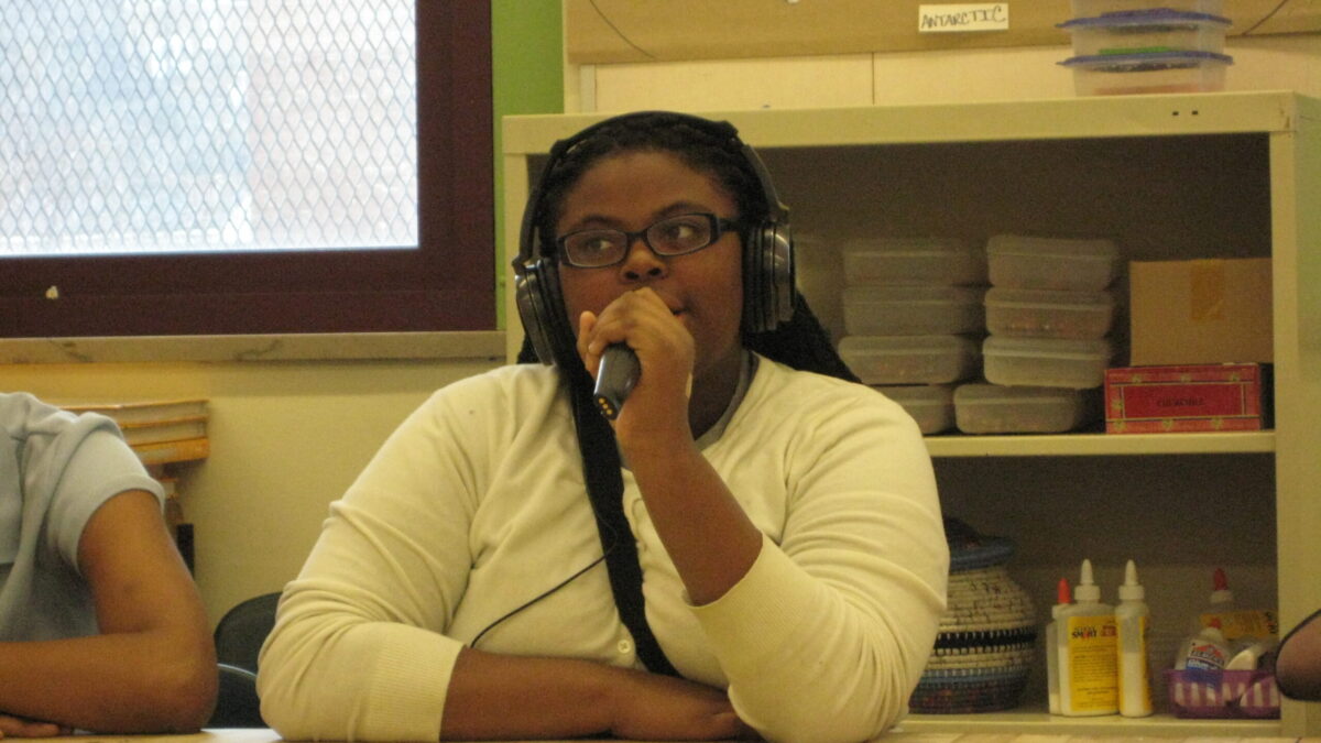 A youth wearing headphones and speaking into a held microphone in a classroom