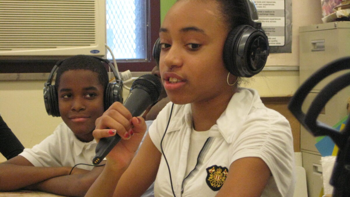 A youth wearing headphones looking at another youth wearing headphones speaking into a held microphone in a classroom.