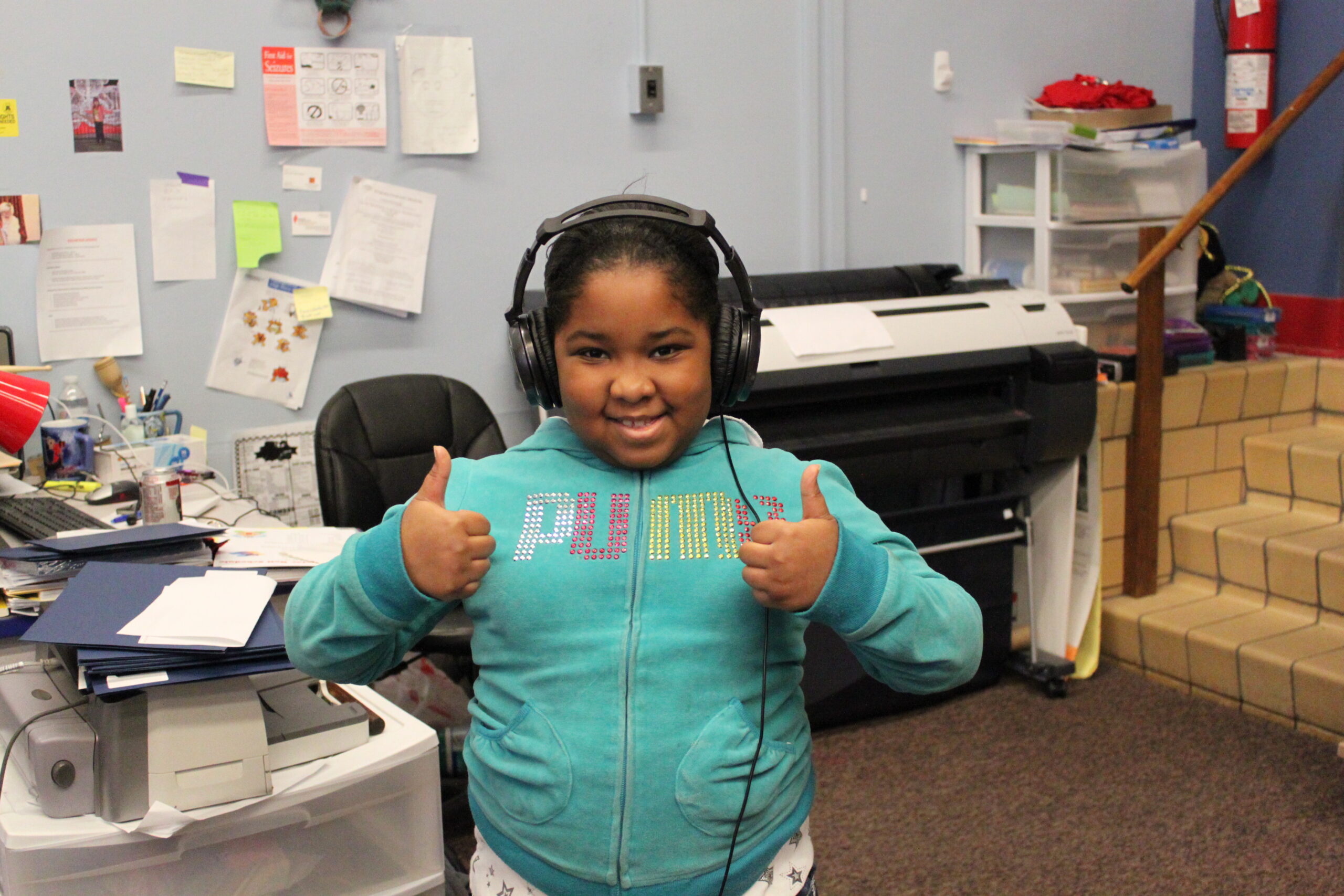 A youth wearing headphones giving two thumbs up at the camera in a classroom