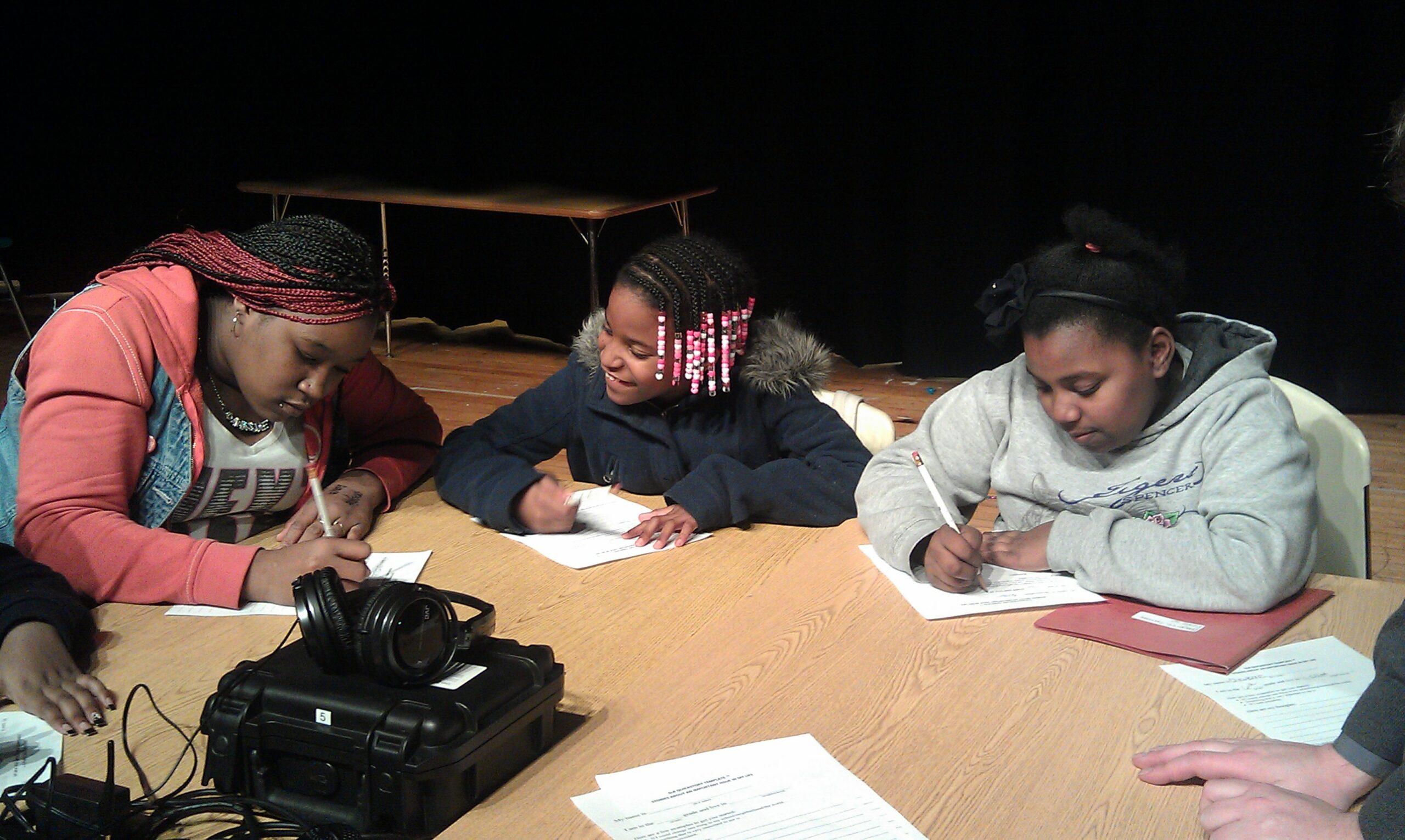 Three youth writing on pieces of paper sitting around recording equipment on a table
