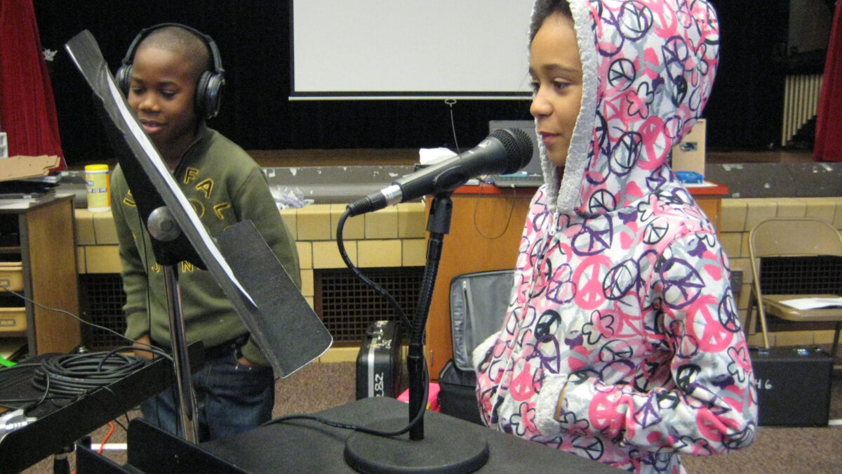 A youth wearing headphones listening to a youth speaking into a microphone while looking at a music stand