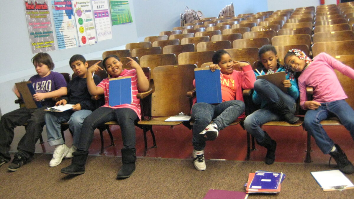 Six youth holding clipboards and notebooks smiling and posing in auditorium seats