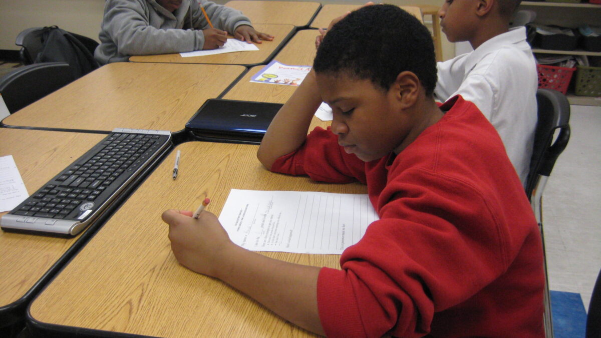 Three youth writing on pieces of paper while sitting at classroom desks