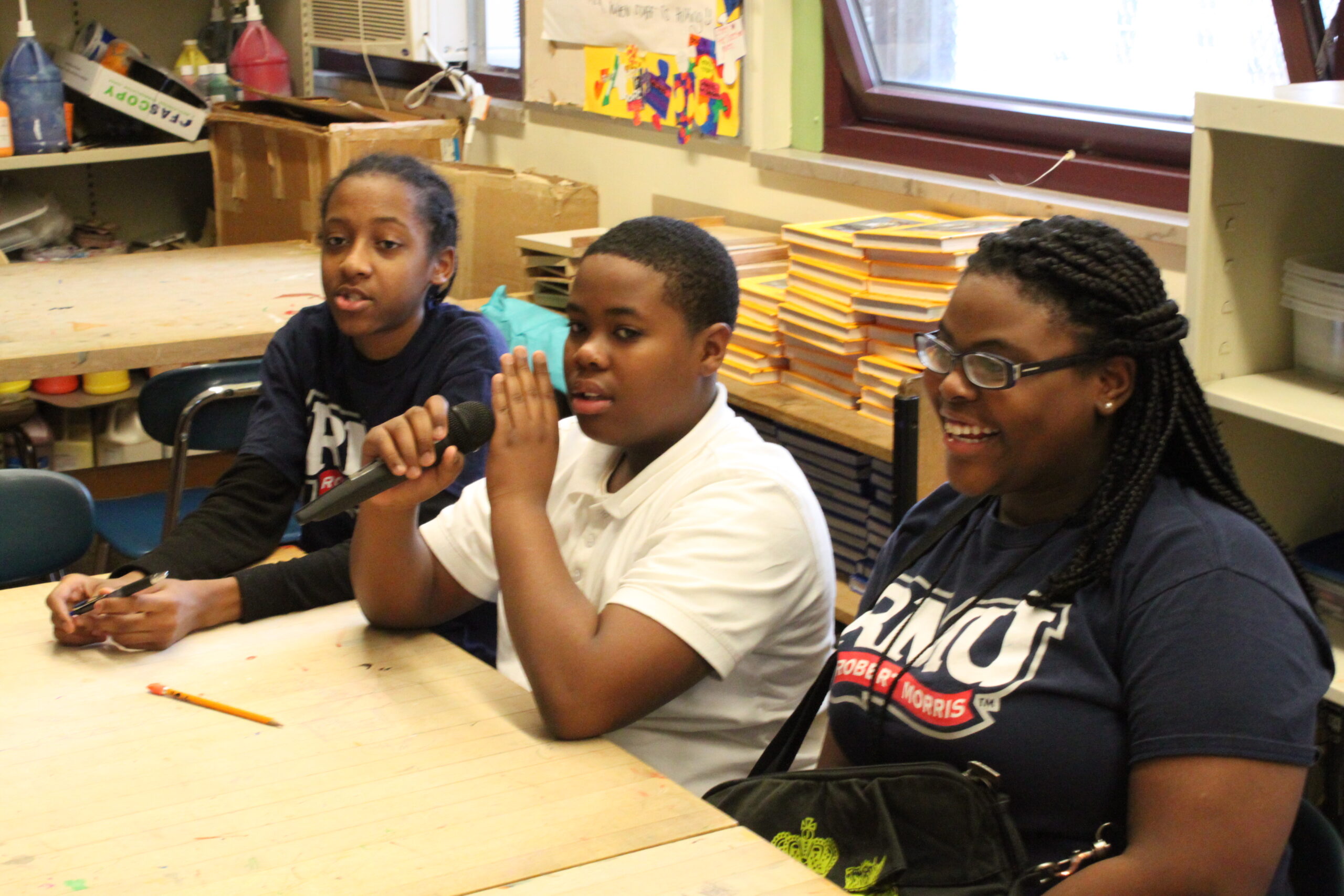 Three youth sitting in a classroom, one holding and speaking into a microphone