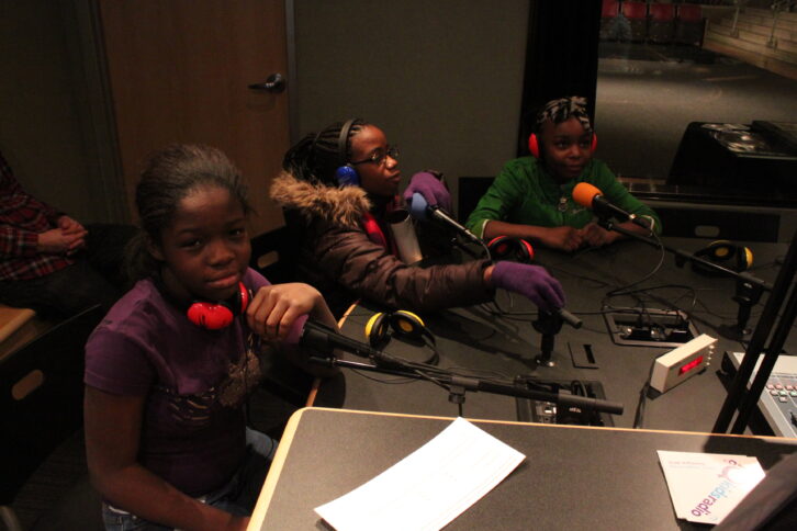Three youth wearing headphones sitting behind microphones in a radio studio.