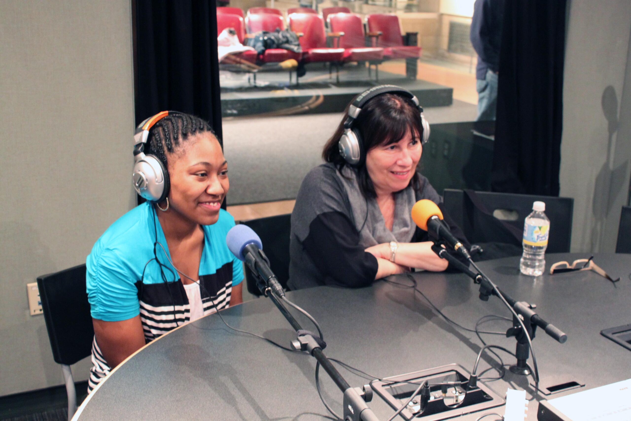 An adult and a teen wearing headphones speaking into microphones in a radio studio