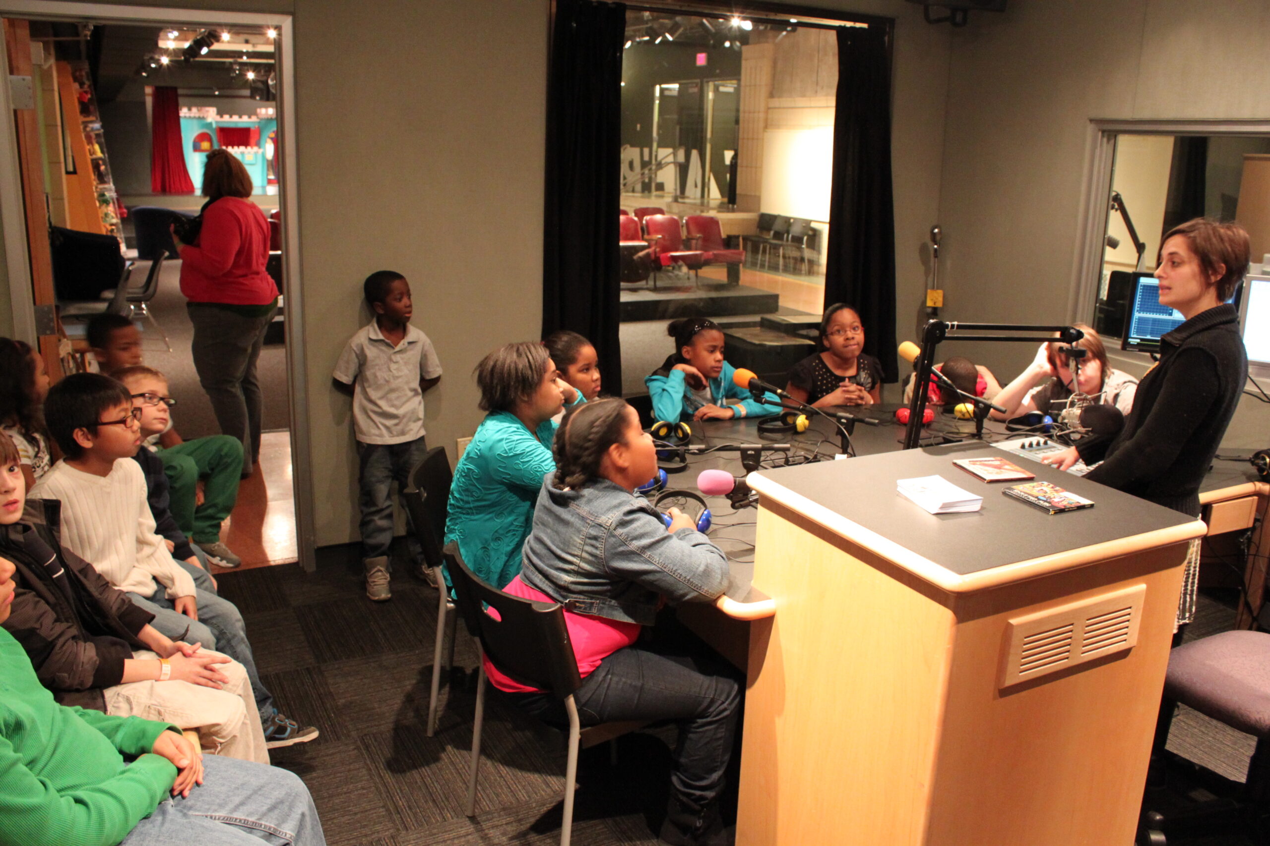 Fourteen youth, seven off to the side, seven sitting behind microphones, listening to an adult talk in a radio studio