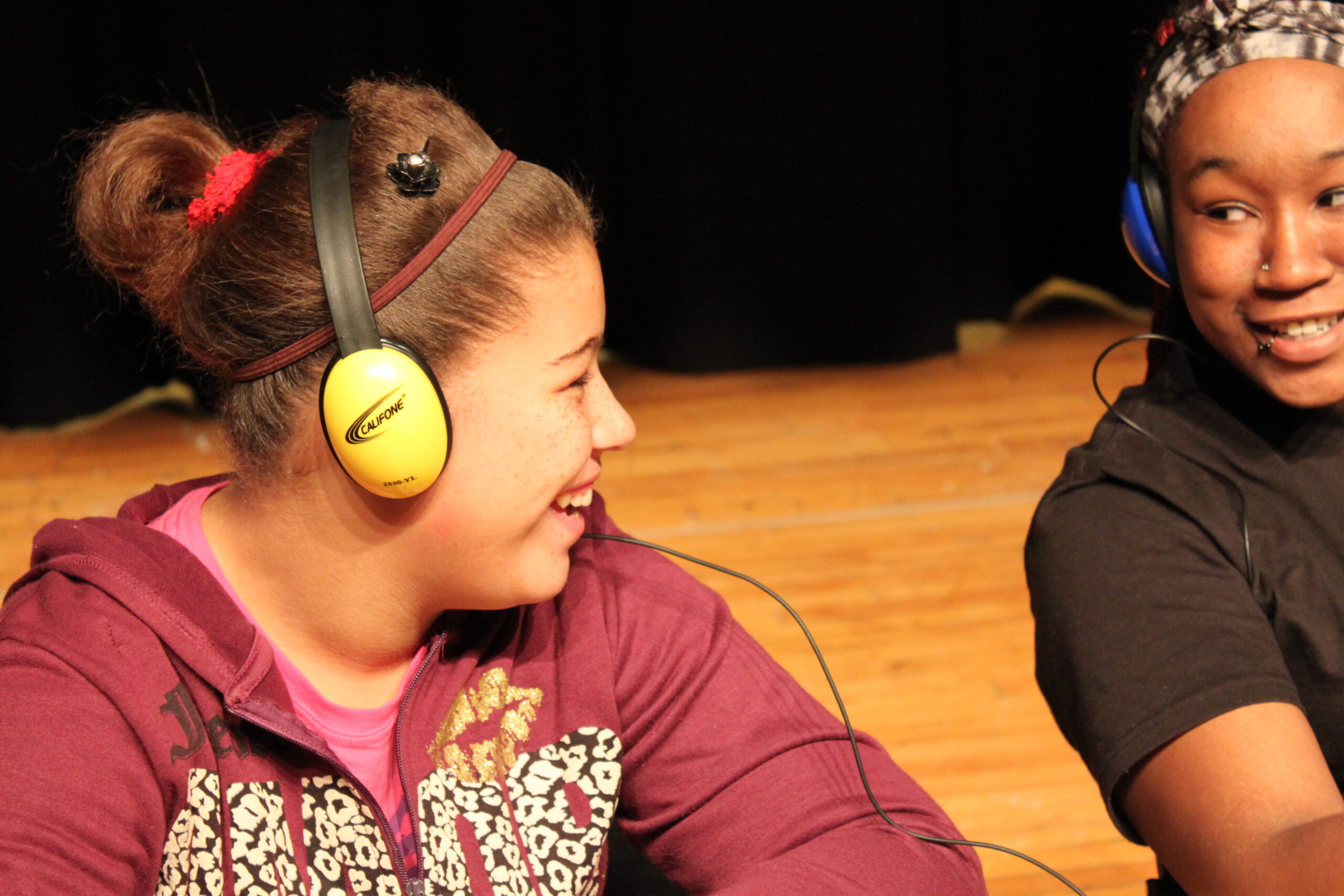 A youth wearing headphones smiling and looking at another youth wearing headphones