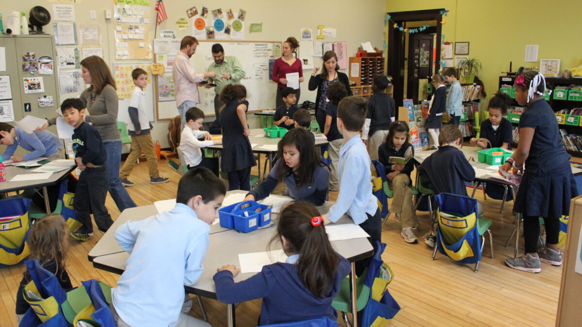 Eighteen youth, eleven sitting at classroom desks, seven standing around the desks, and five adults overseeing while standing in front of a whiteboard.